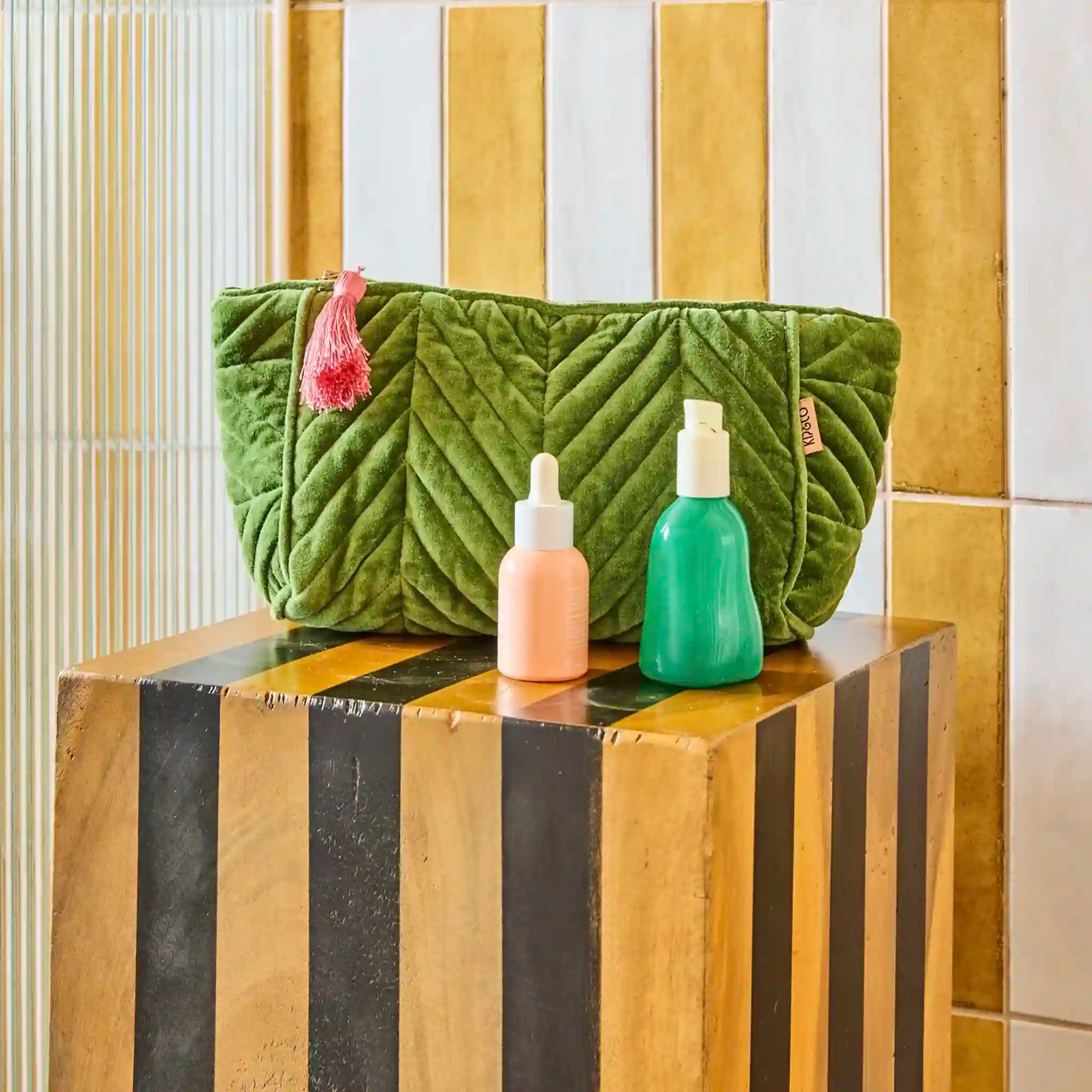Green textured toiletry bag with a pink tassel on a wooden block against a striped wall.