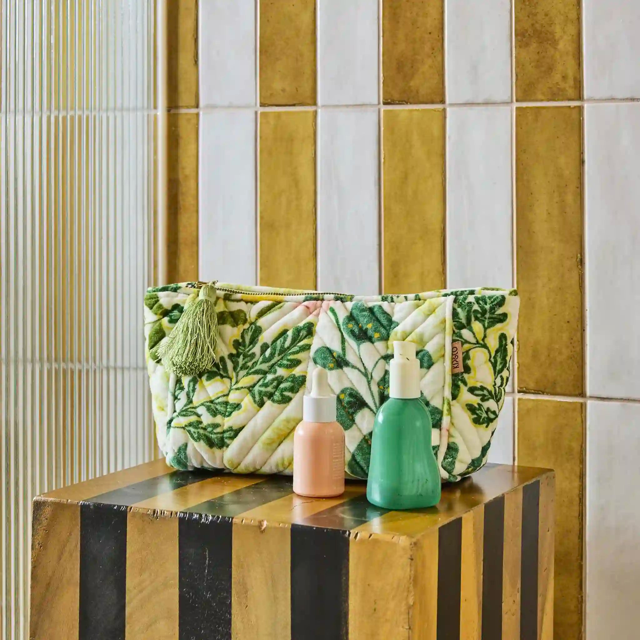 Green and white patterned toiletry bag with two bottles on a striped surface against a tiled wall.