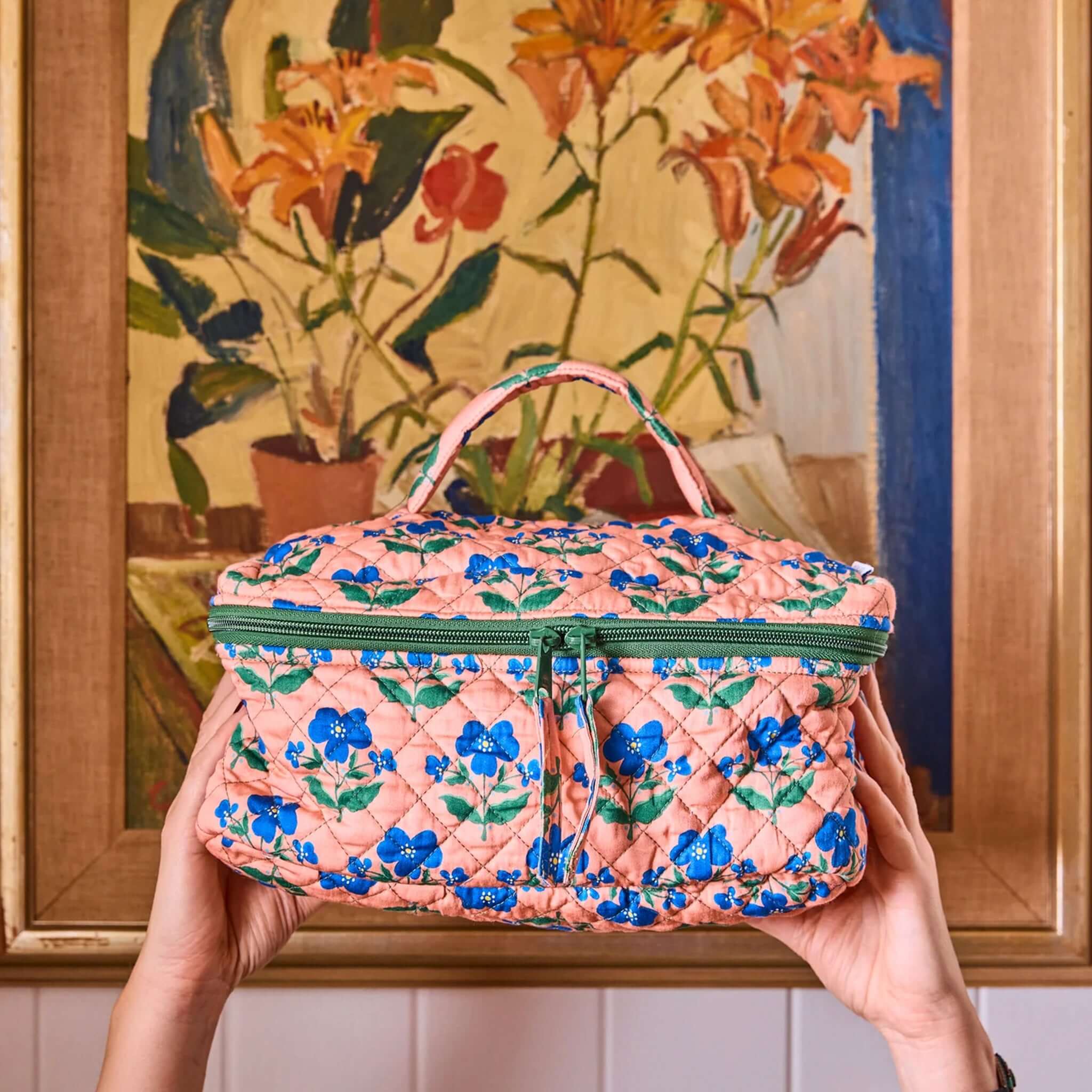 Kip & Co x Anna Spiro Quilted Toiletry Case - Indian Summer Rose held in front of a floral painting.