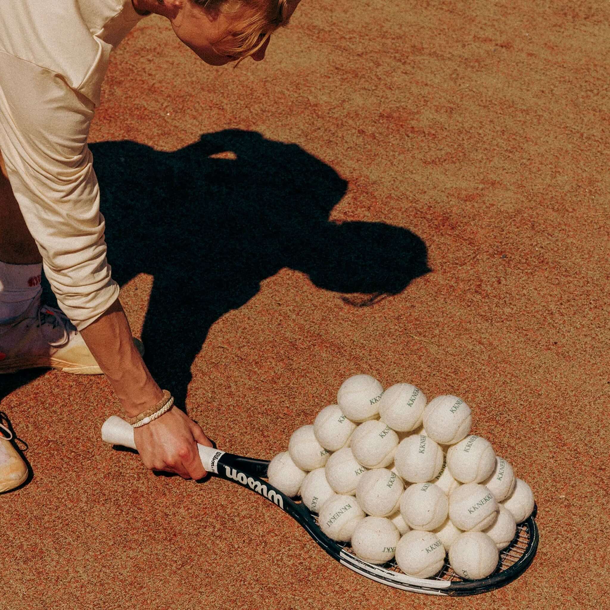 A hand holding a tennis racket with a pyramid of tennis balls on a clay court.