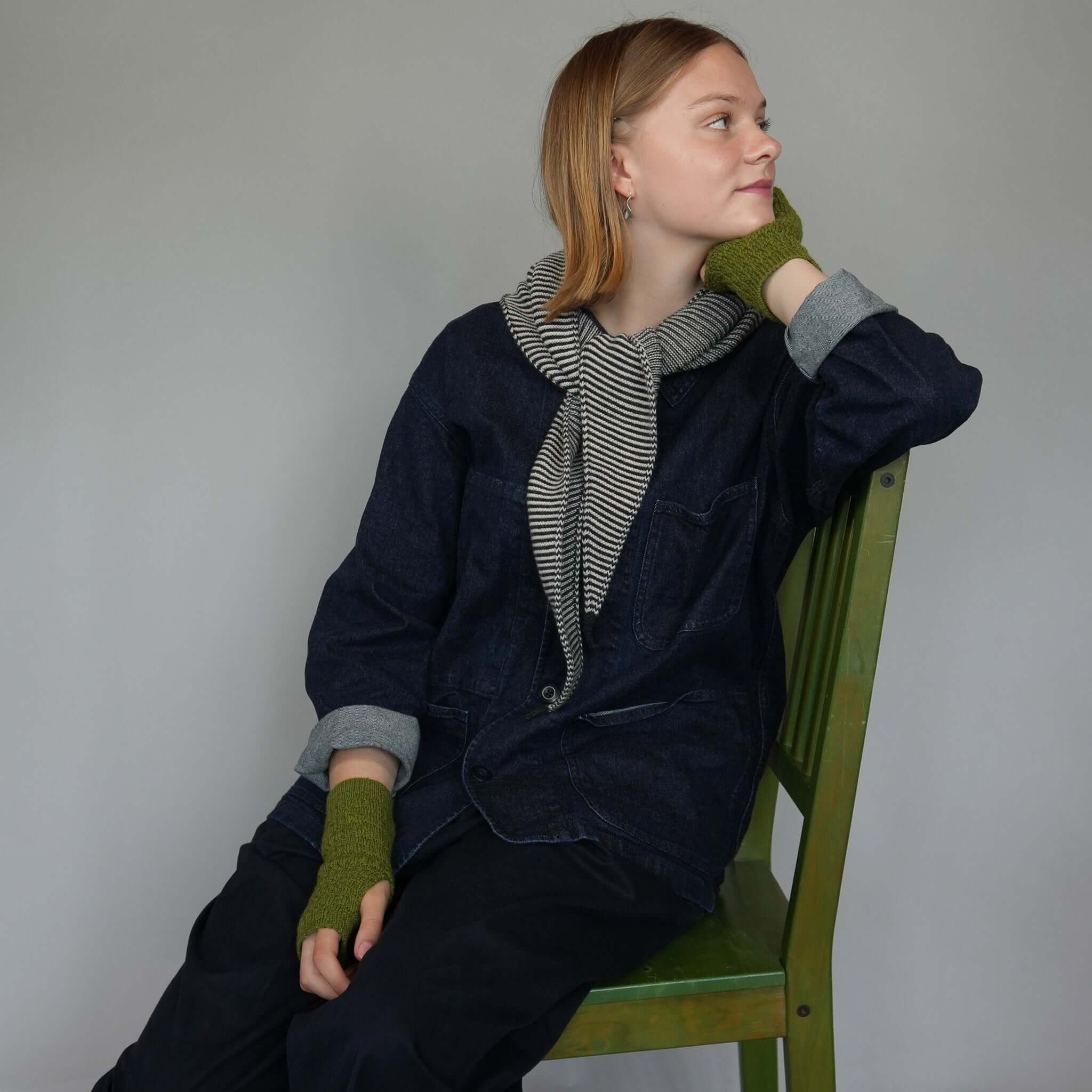 Model wearing Knit Shepherd Punakaiki Merino Neckerchief - Ponga Black, seated on a green chair.
