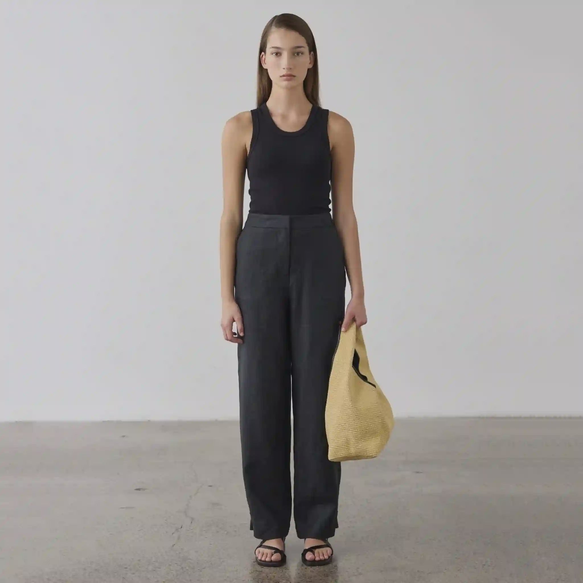 Woman in black sleeveless top and pants holding a yellow bag against a plain background