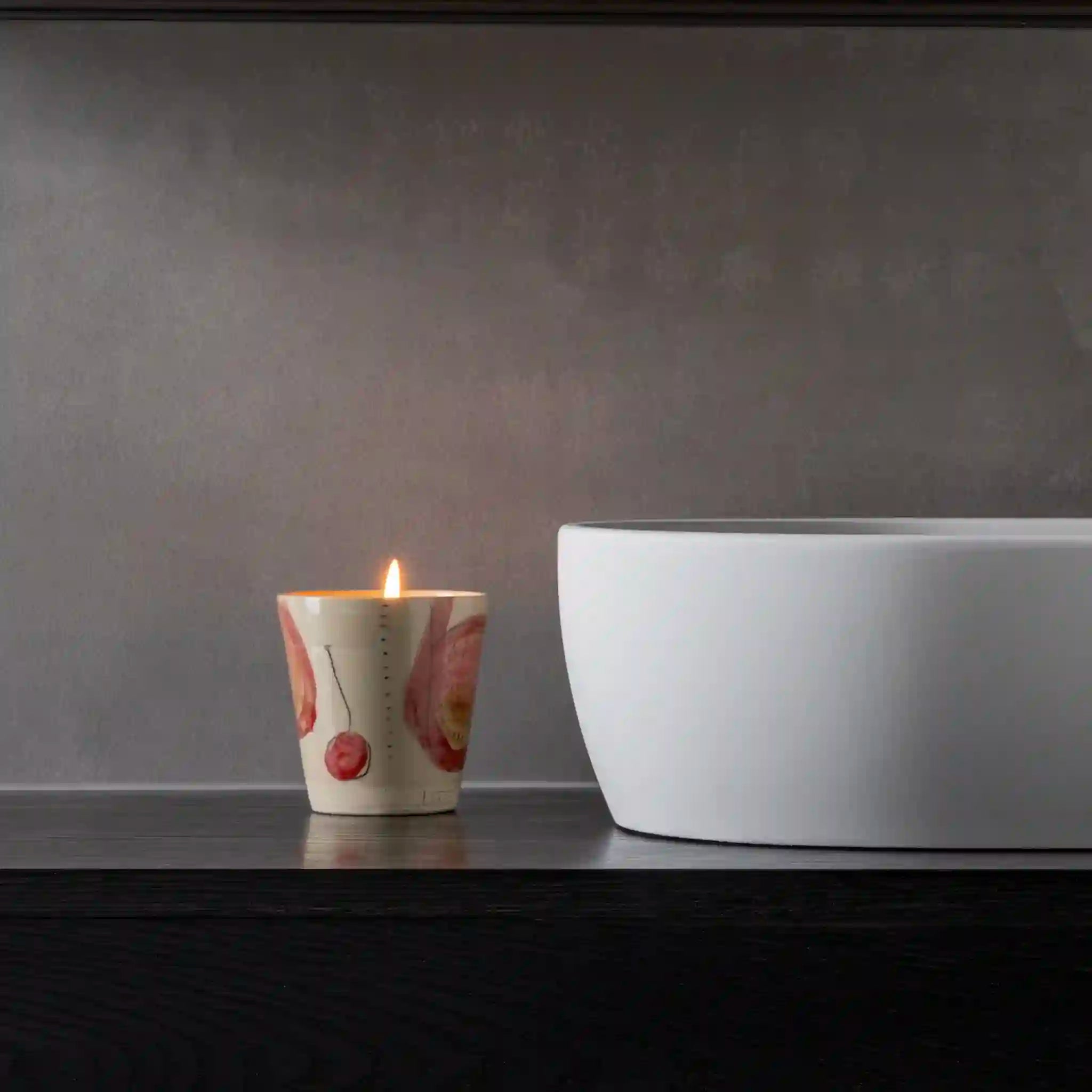 Candle with a floral design next to a white bathtub against a gray wall.