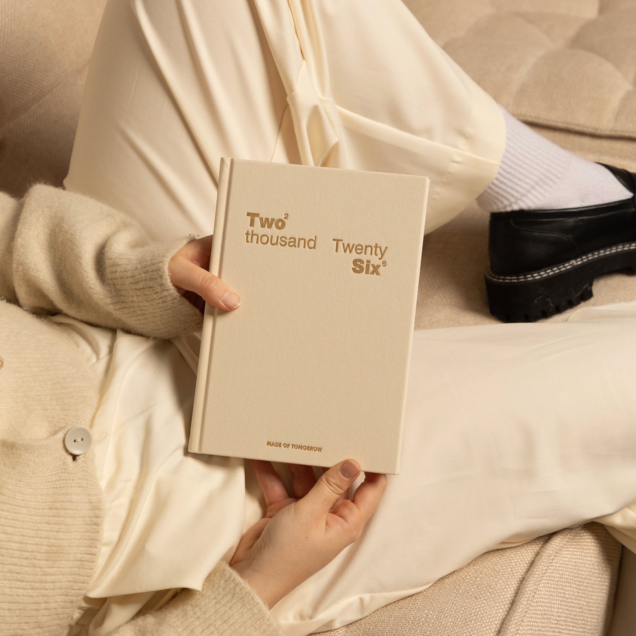 Person holding a beige book titled 'Two thousand Twenty Six' on a couch.