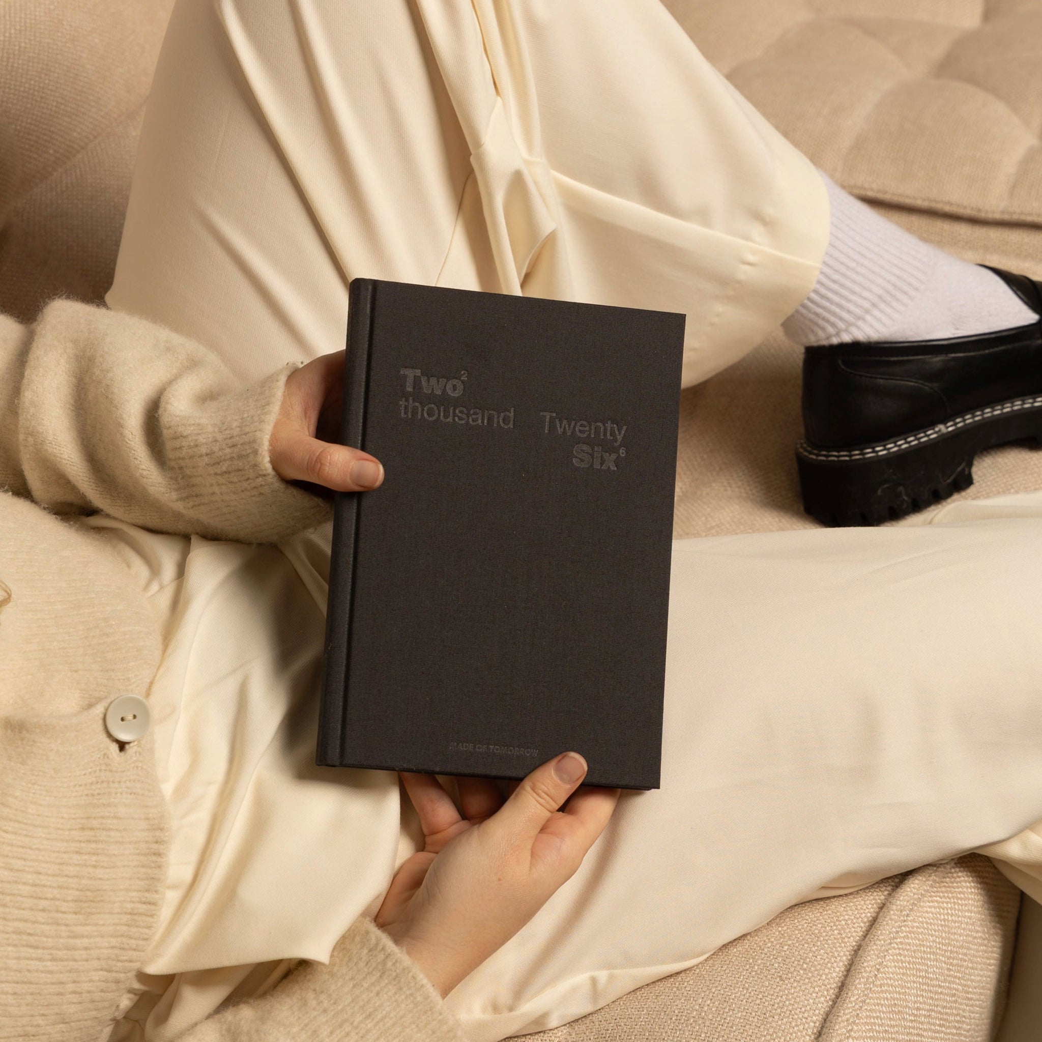 Person holding a black book titled 'Two thousand Twenty Six' on a beige couch.