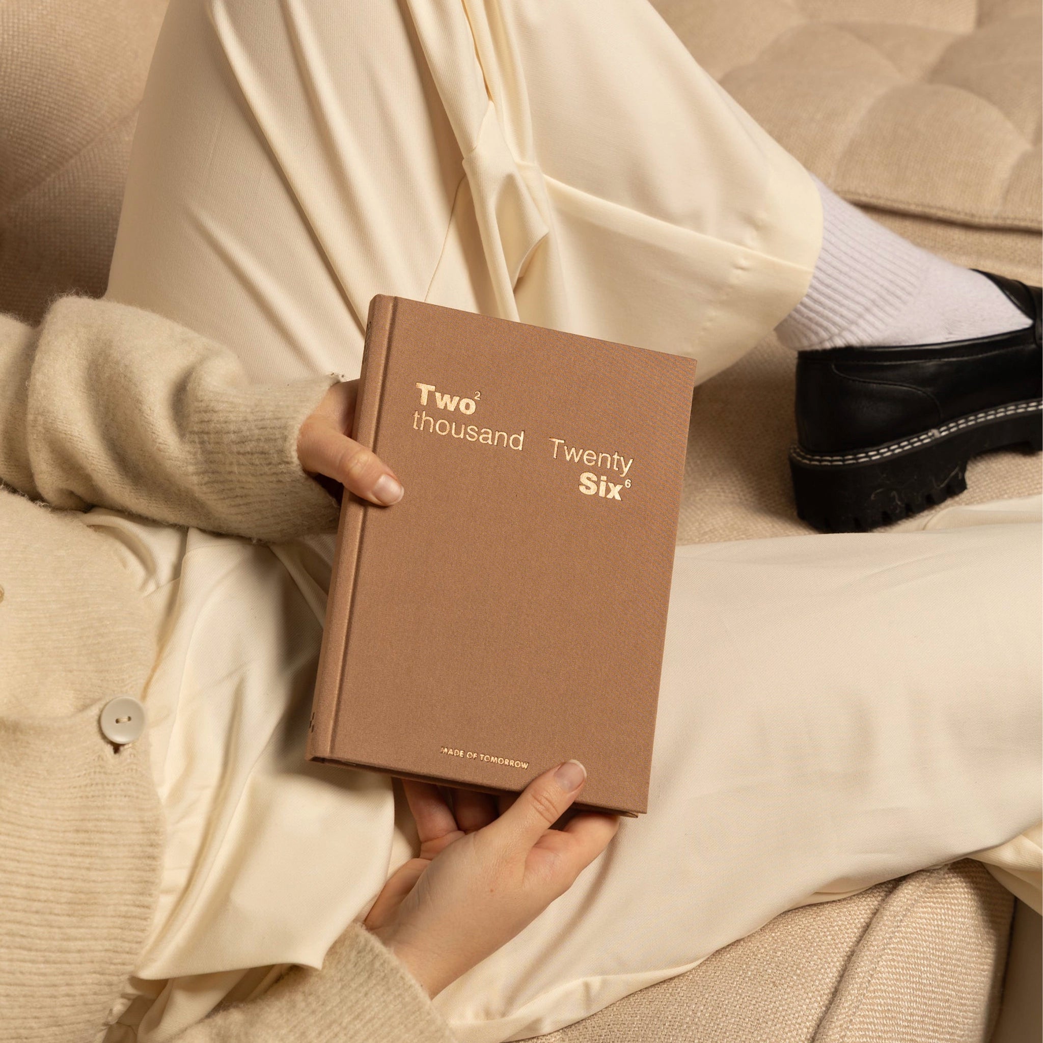 Person holding a brown book titled 'Two thousand Twenty Six' on a beige couch.