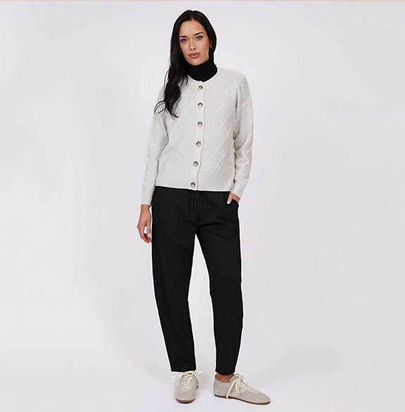A woman modeling a light beige cable-knit cardigan with gold buttons over a black turtleneck, paired with black pants and beige sneakers, against a plain white studio background.