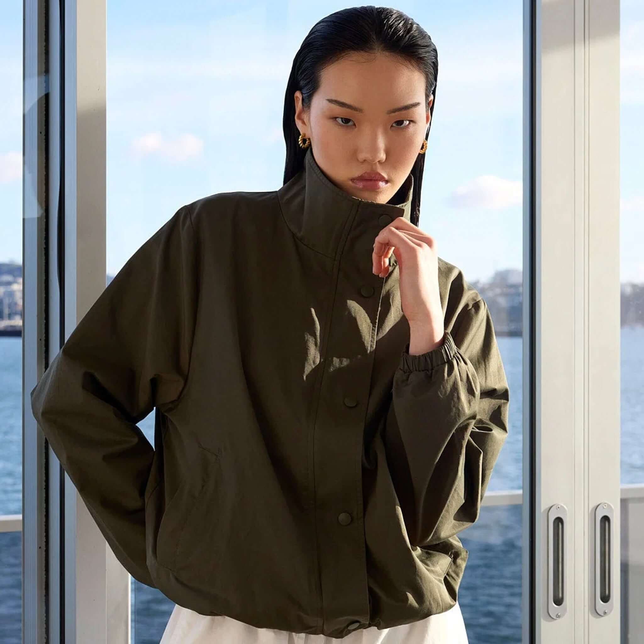 Model wearing Marlow Rift Jacket - Olive with a funnel neck, showcasing casual street-style in front of a scenic view.