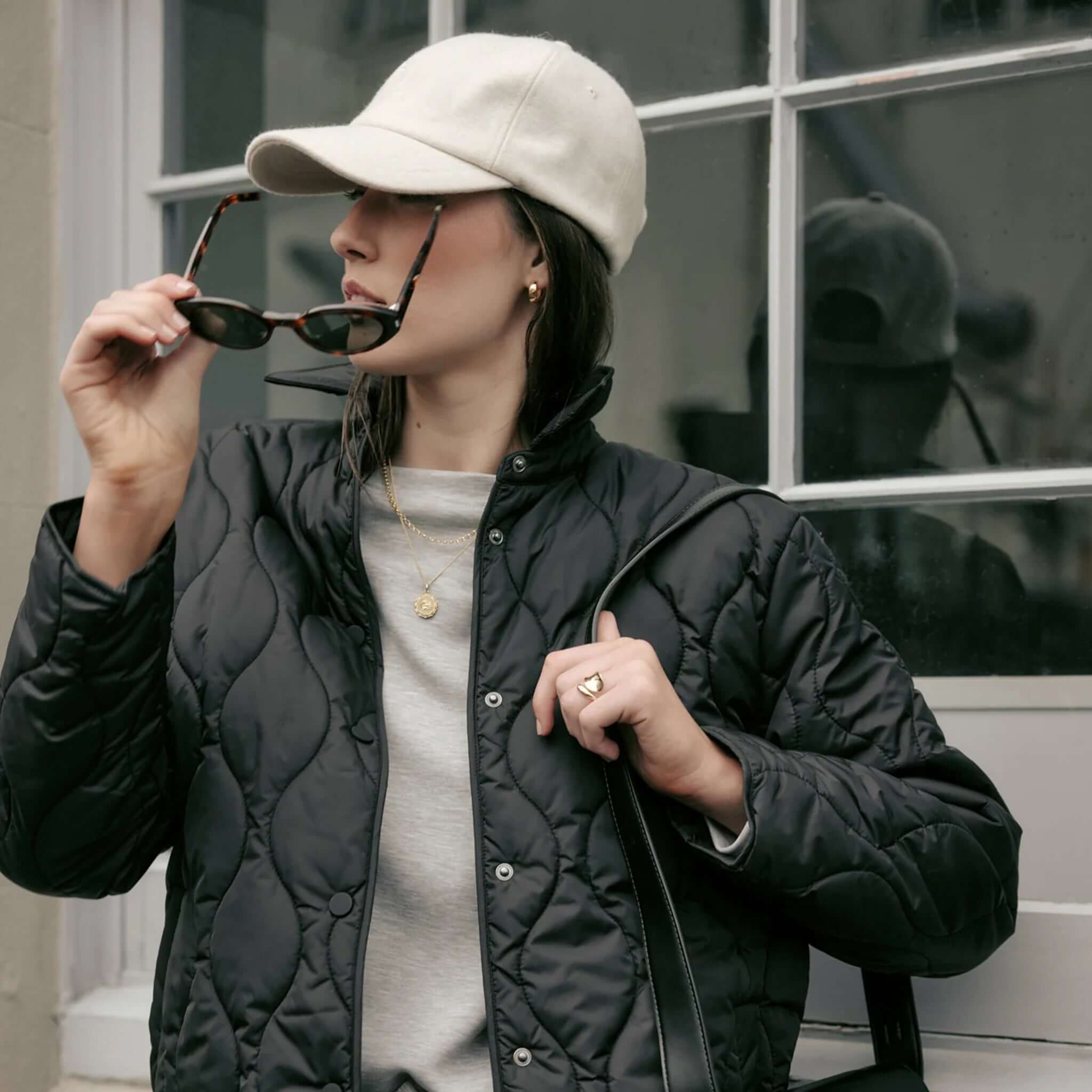 Marlow Toronto Quilted Jacket - Black worn by a person holding sunglasses and wearing a baseball cap.