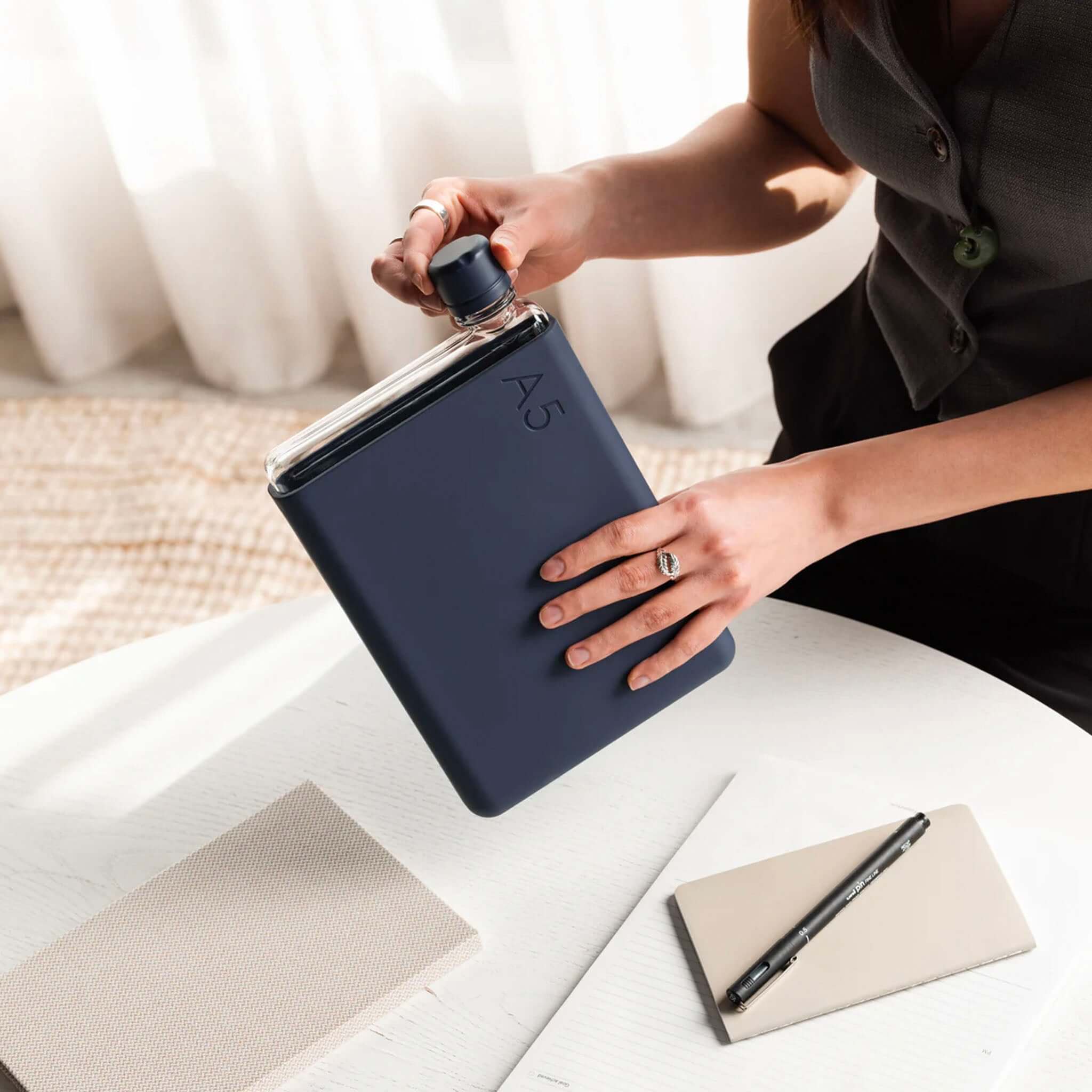 Alt Tag: Memobottle Bundle A5 - Black Ink with matching silicone sleeve and lid, flat pocket water bottle on desk beside notebook and pen