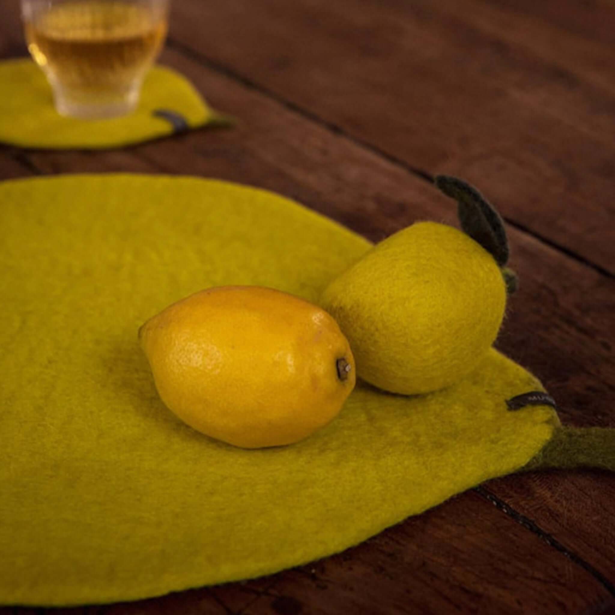 Muskhane Felt Lemon Placemat - Sulfur Flower yellow felt lemon-shaped placemat on wooden table with lemon and glass