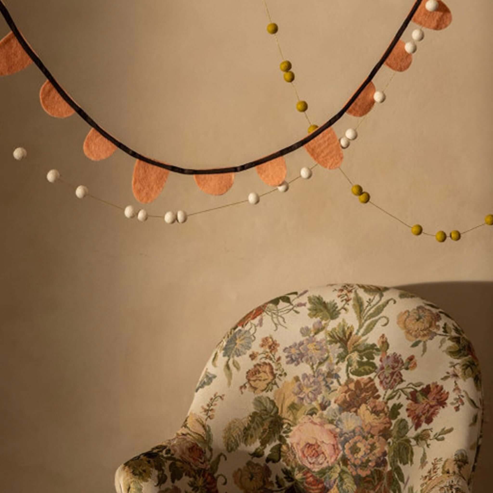 Muskhane Felt Round Flag Garland - Blush draped elegantly against a wall with a decorated chair.