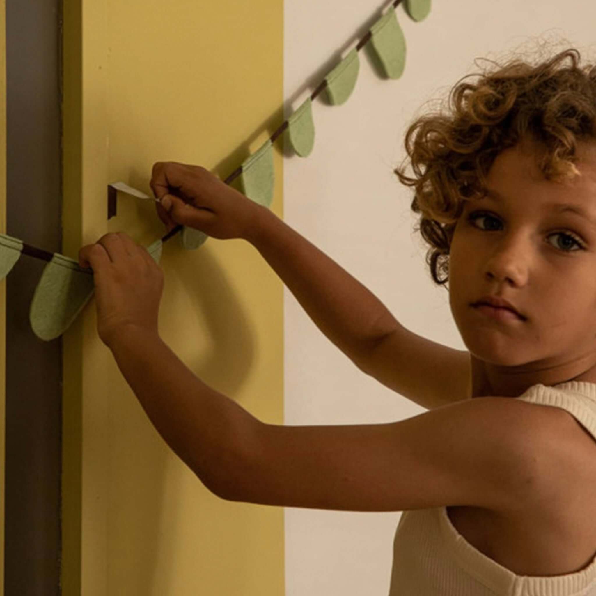 Child hanging Muskhane Felt Round Flag Garland - Mint on a wall, showcasing its playful and colorful design.