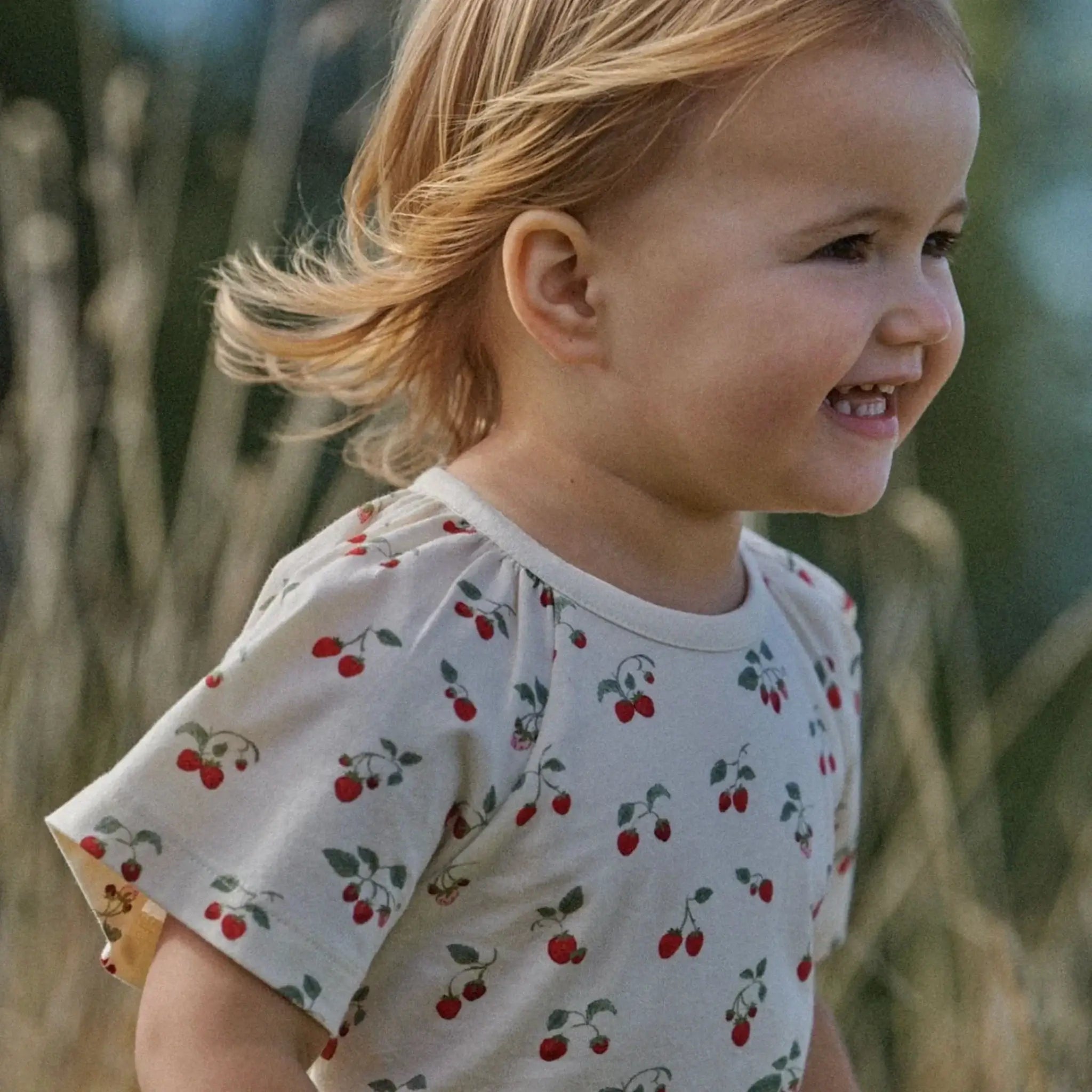 Alt Tag: Nature Baby Organic Cotton Faye Tee - Strawberry Blush toddler wearing organic cotton tee with gathered sleeves and cherry print outdoors
