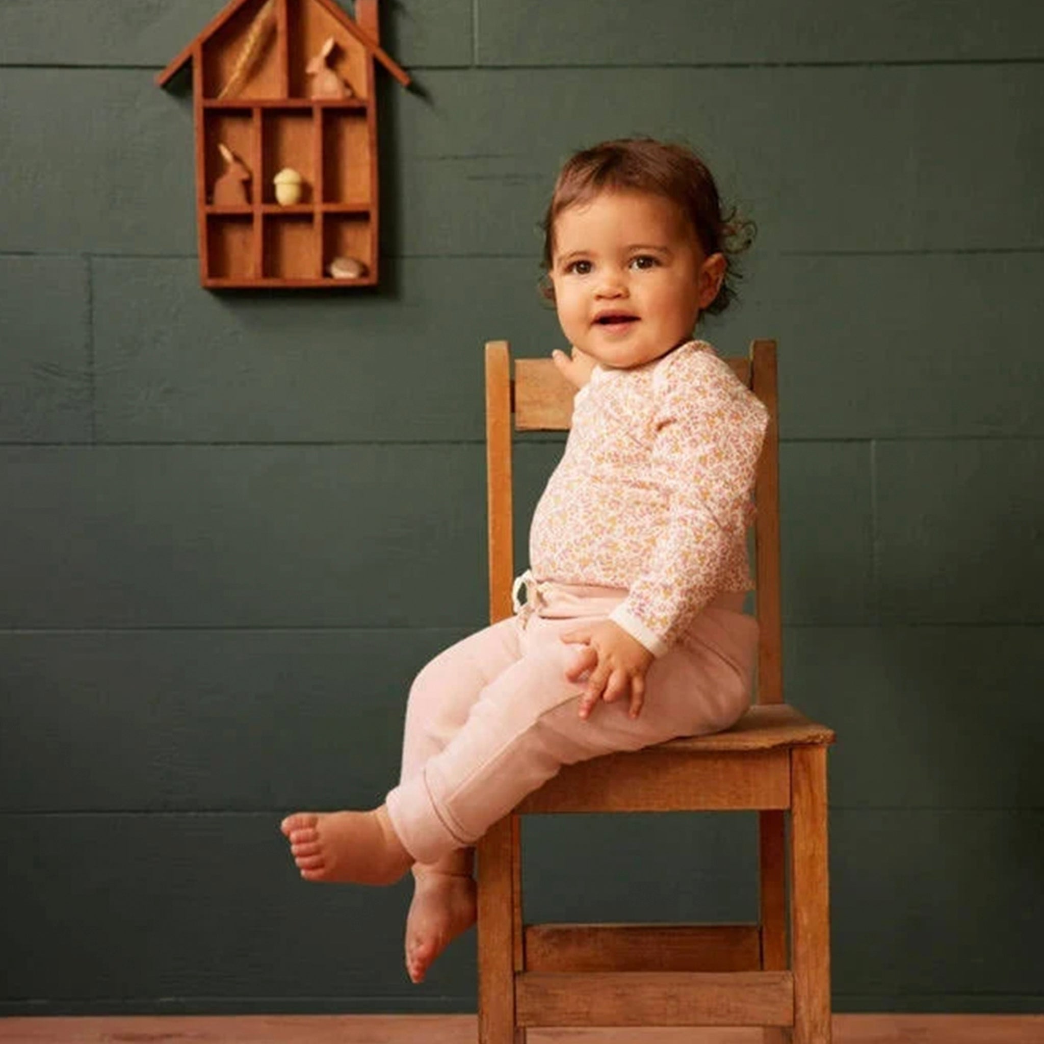 Child sitting on a wooden chair against a green wall with a small wooden shelf.