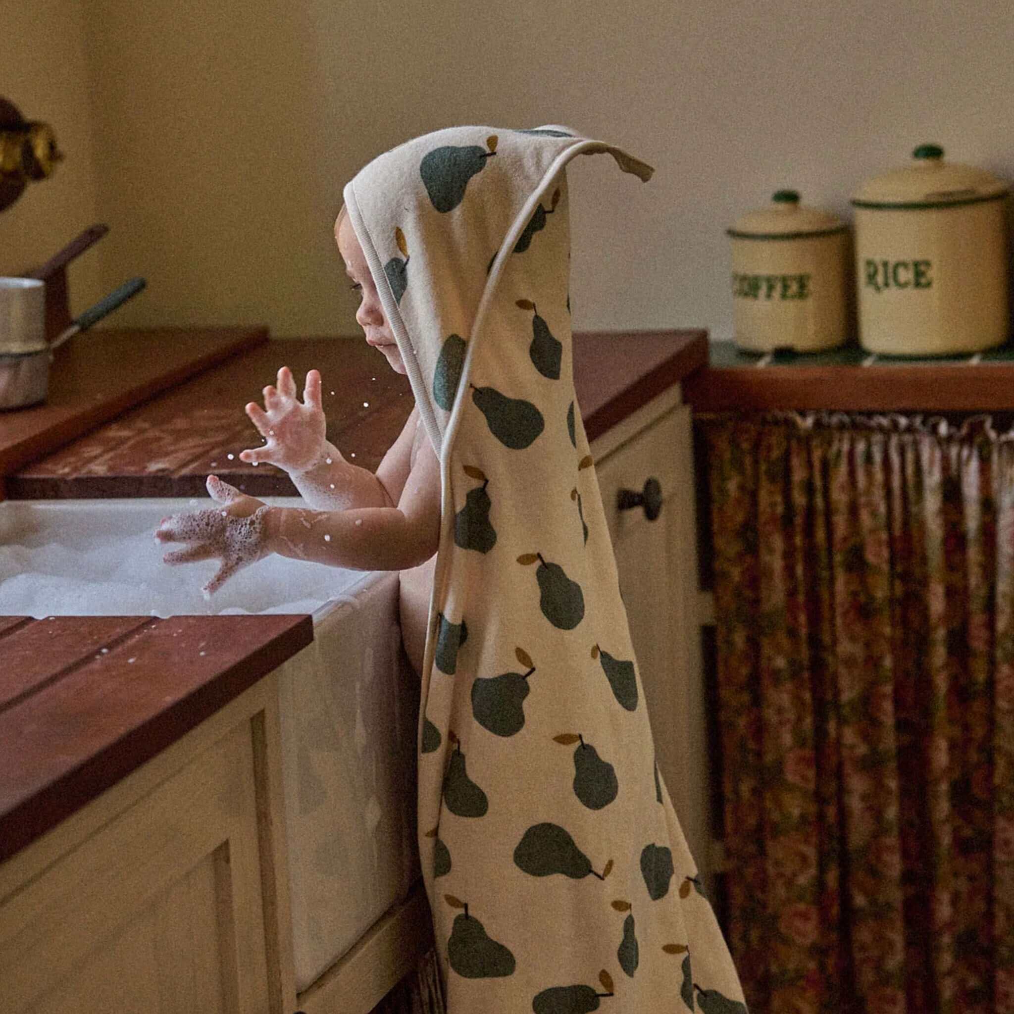 A toddler wearing a Nature Baby Organic Cotton Hooded Towel - Pear, playing with bubbles at the sink.
