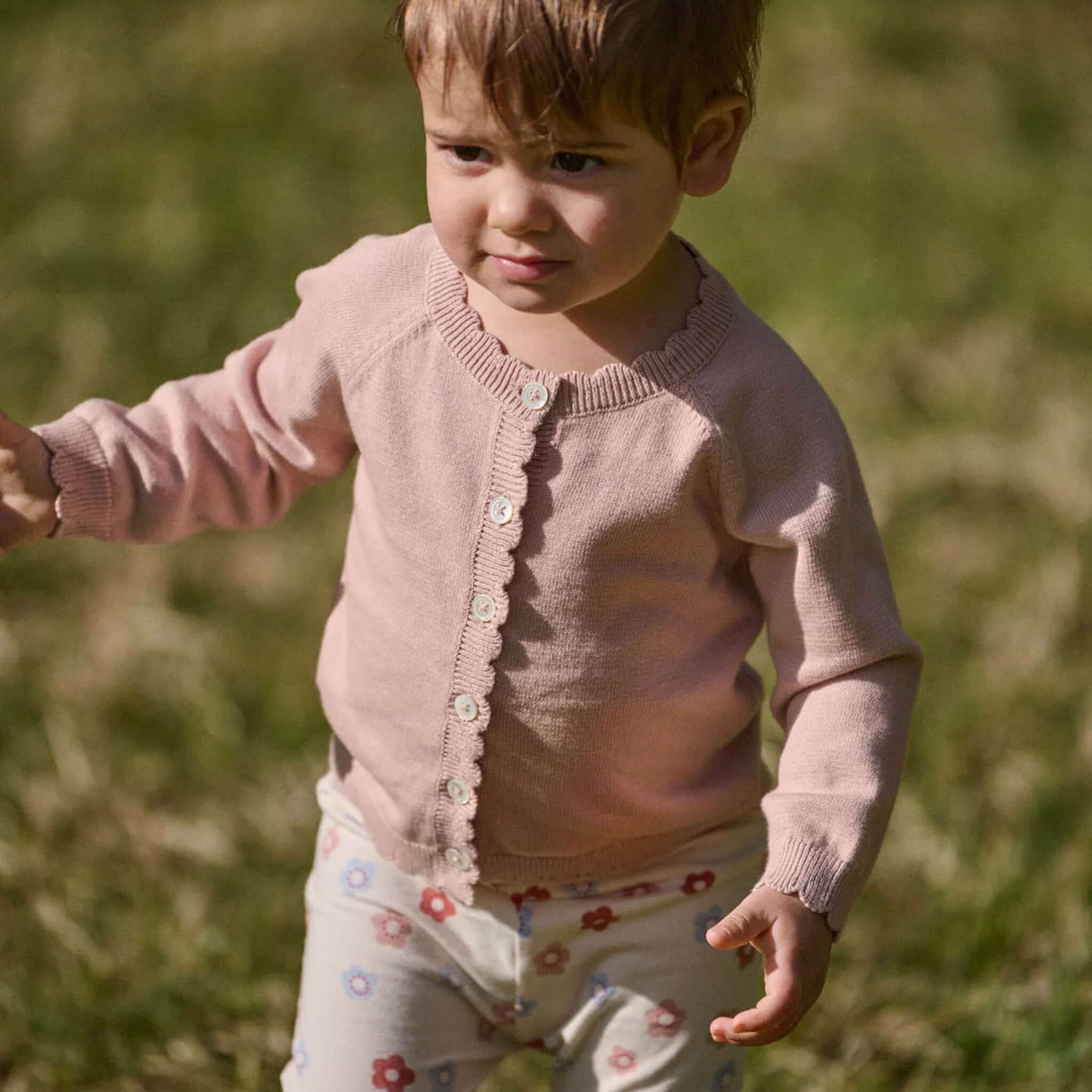 Nature Baby Organic Cotton Liliette Cardigan - Rose Dust worn by a toddler outdoors, showcasing scalloped trim and natural button closures.