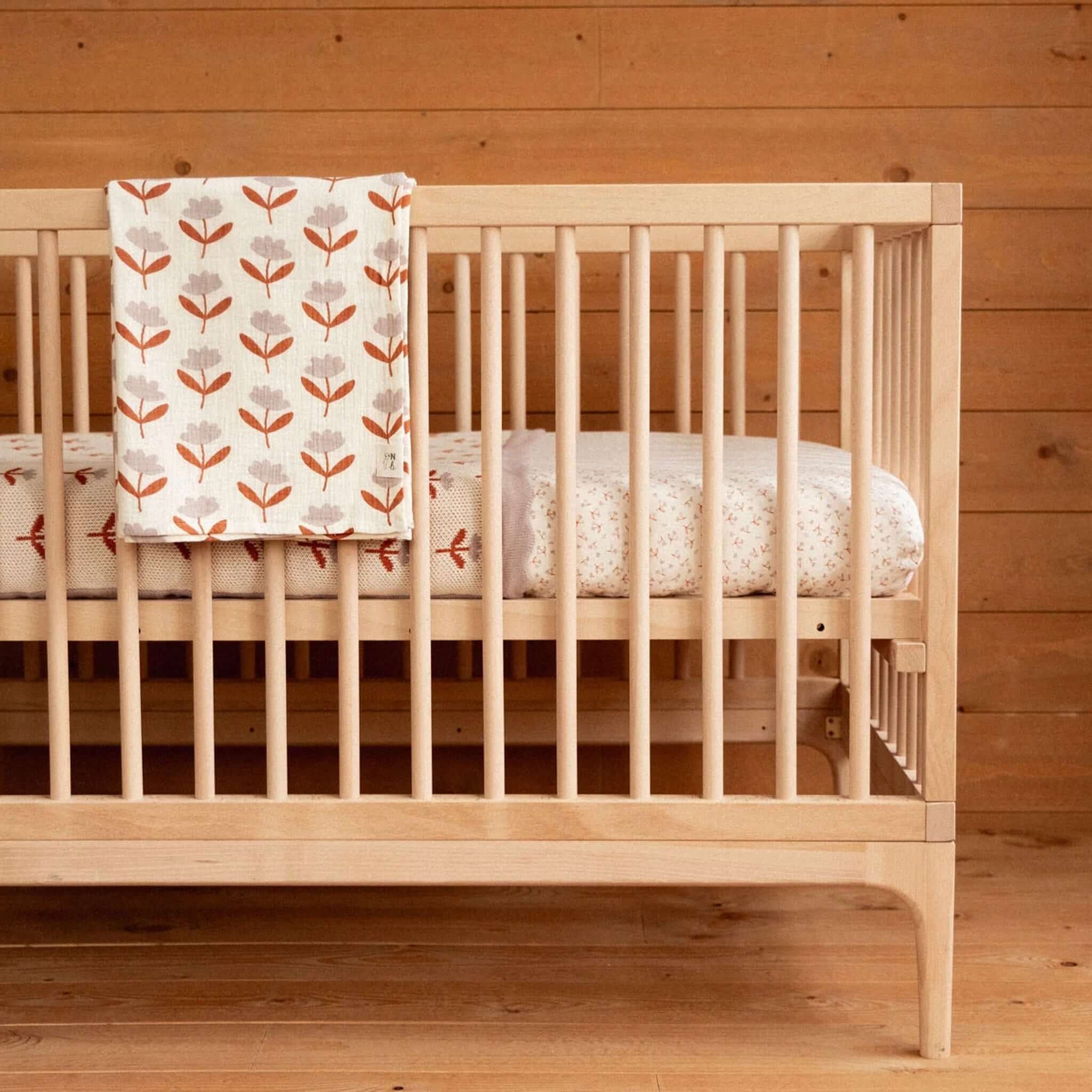 Nature Baby Organic Cotton Muslin Wrap - Fleurette hanging on a wooden crib, showcasing its floral design and soft fabric.