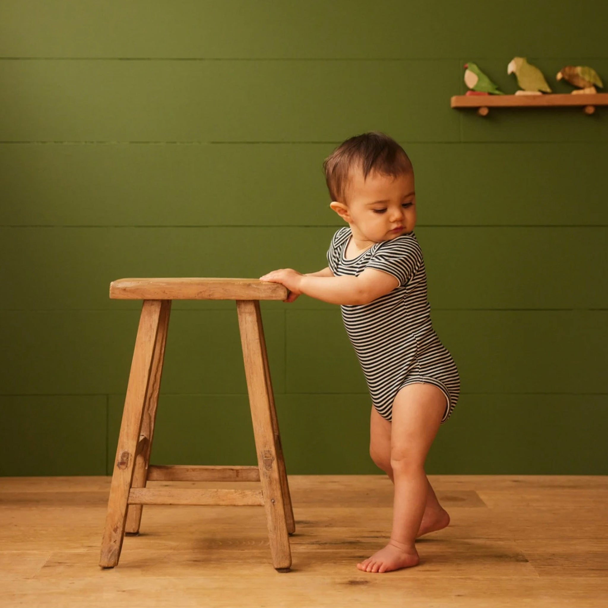 Baby wearing a navy and white striped short sleeved baby bodysuit holding onto a wooden stool.