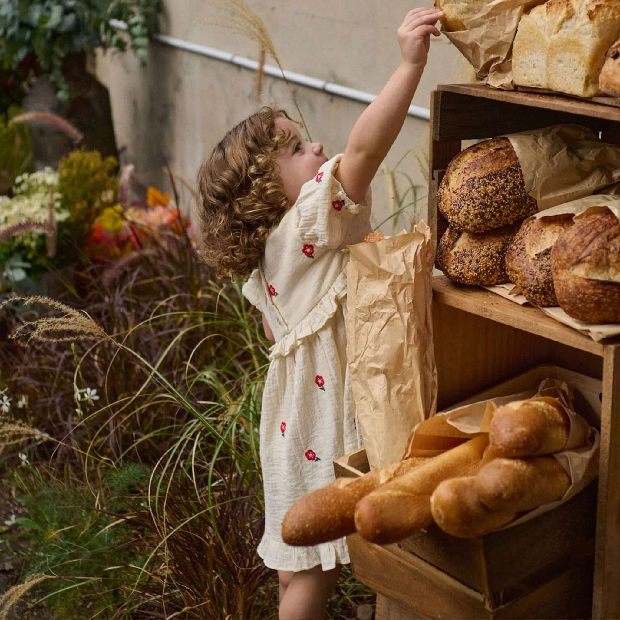Child reaching for fresh bread while wearing Nature Baby Organic Cotton Valentina Dress - Red Flora with floral details.