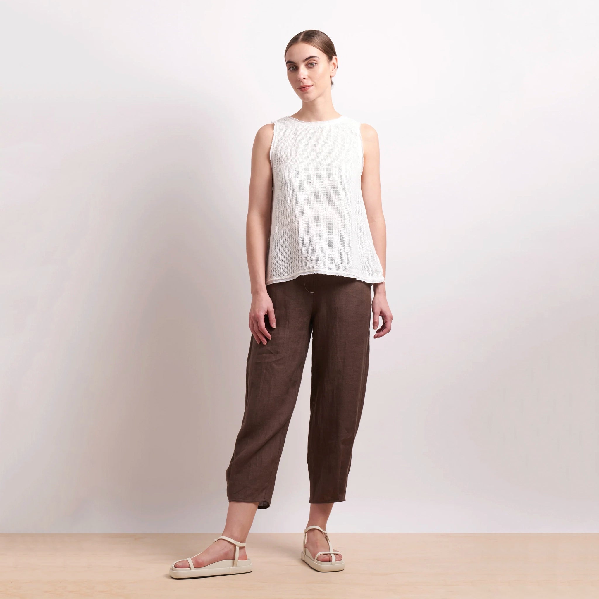 Woman wearing a white sleeveless top and brown pants on a plain background