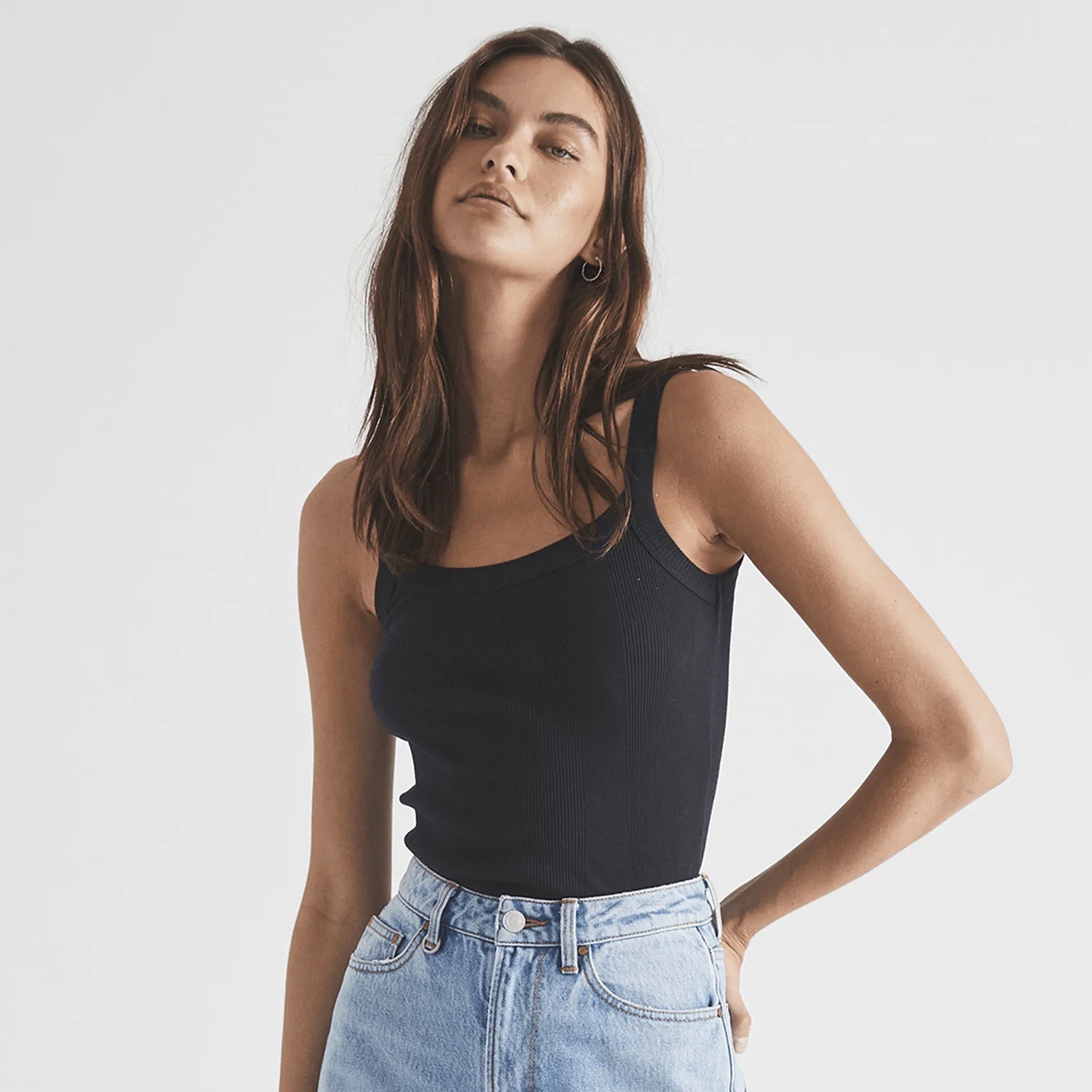 A woman wearing a black organic cotton rib singlet with a slim fit and irregular rib detailing, paired with denim jeans.