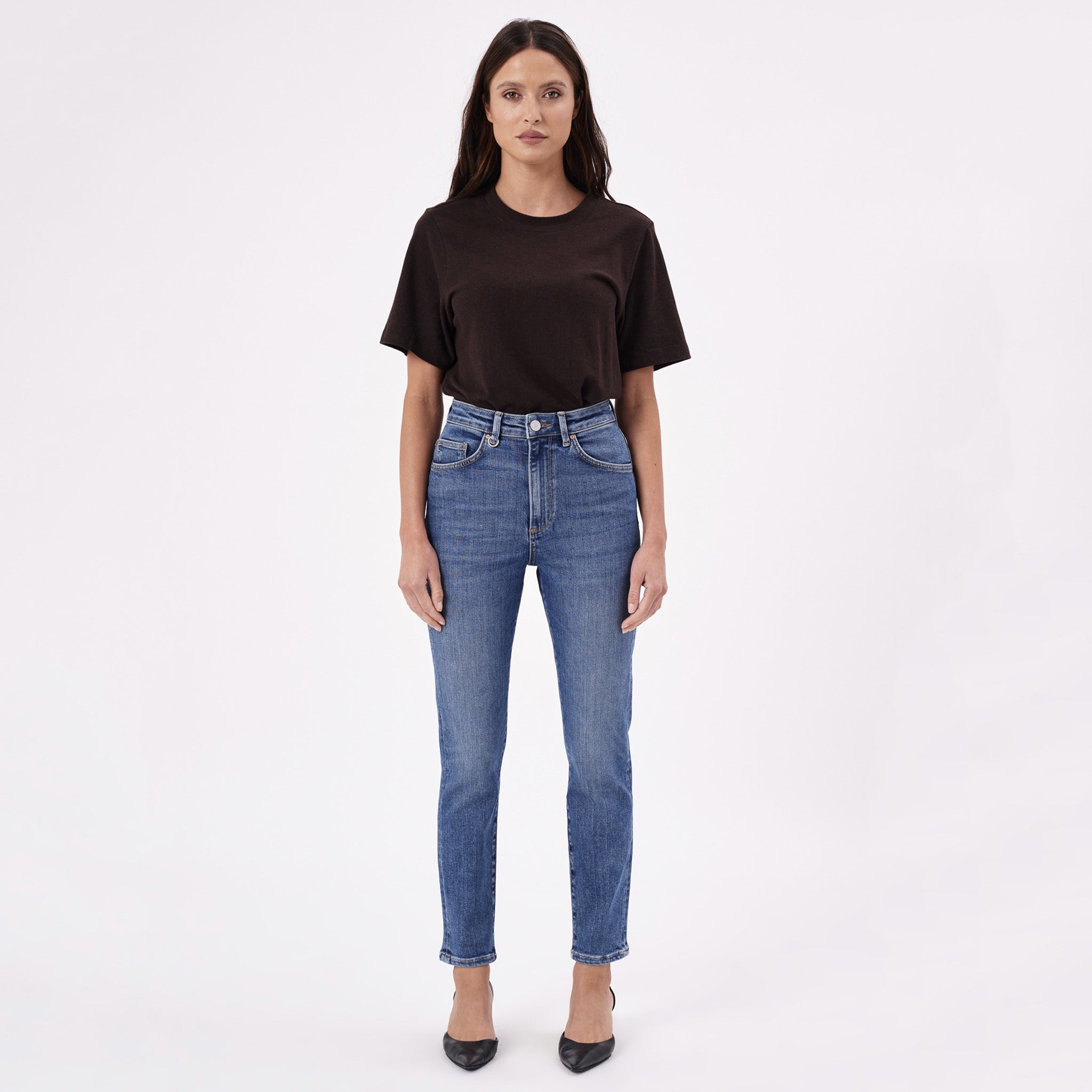A person wearing a brown top and blue jeans, standing against a white background.