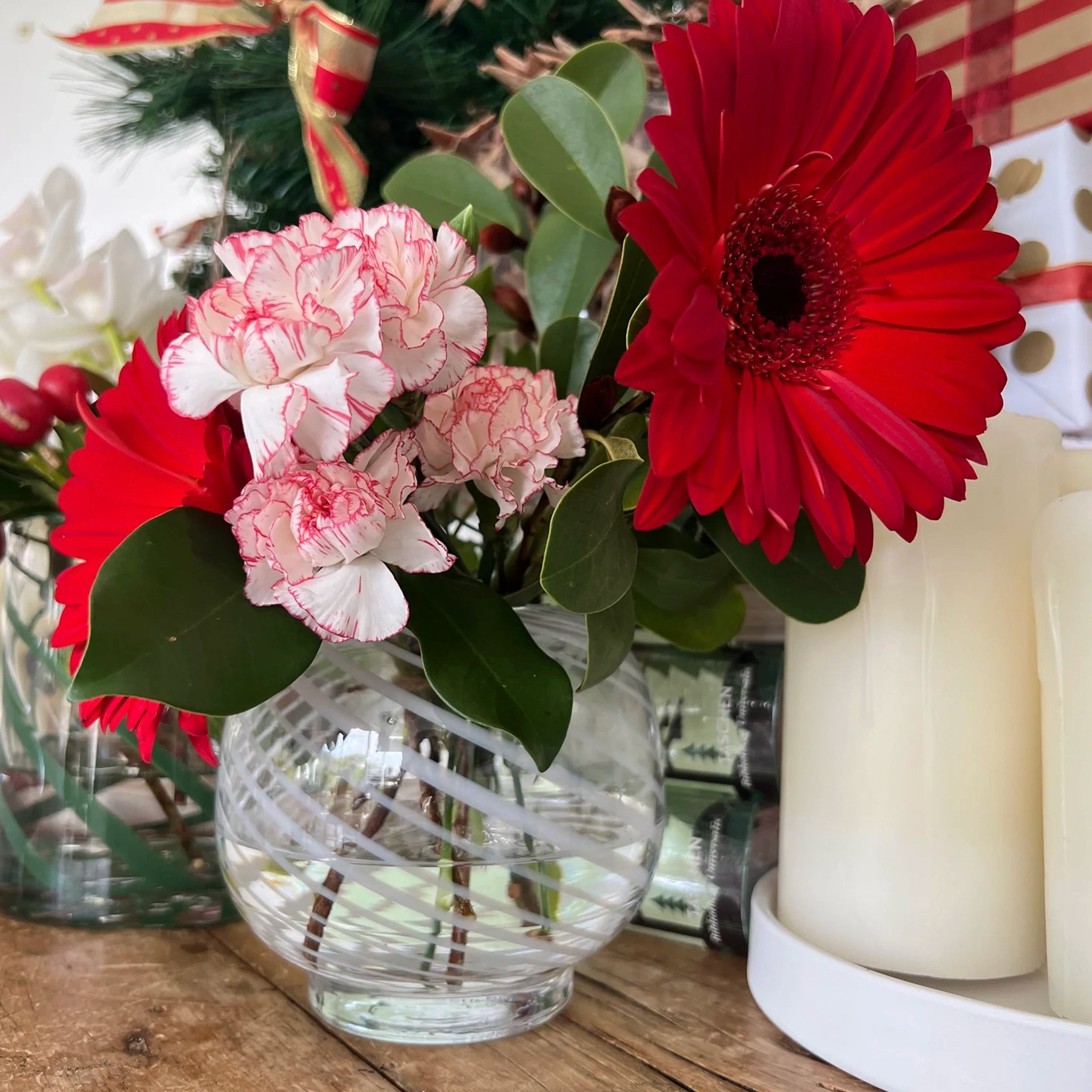 Clear glass vase with red and pink flowers on a wooden surface with candles and greenery.