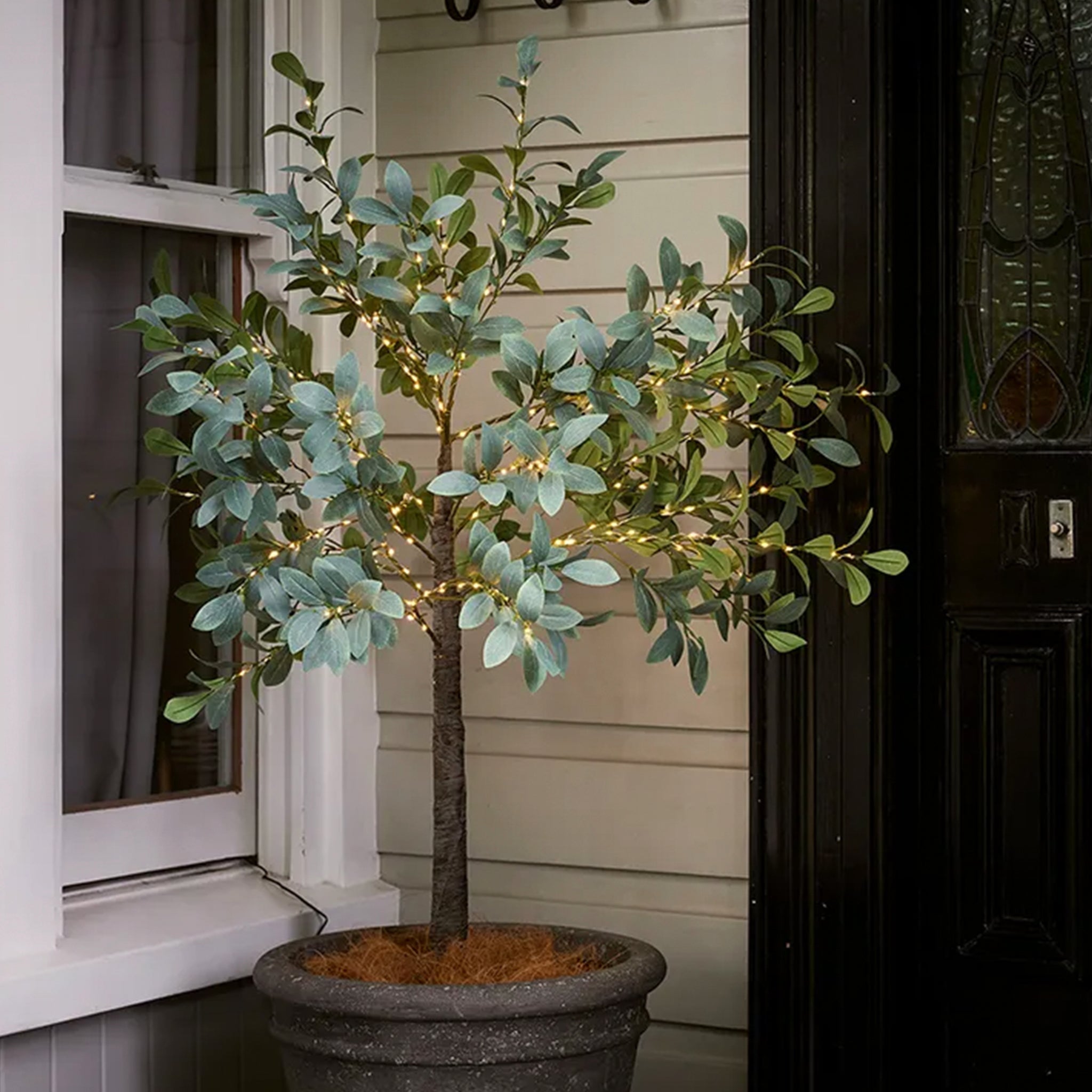 An artificial olive green Christmas tree decorated with warm white LED lights in a pot.