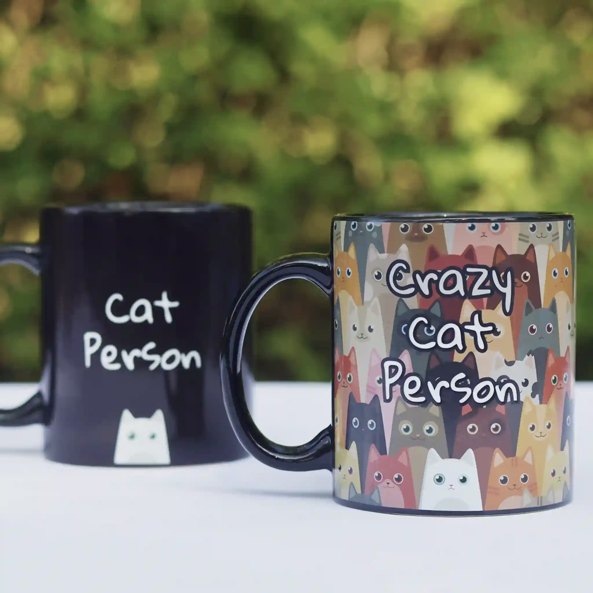 Two mugs; one black with 'Cat Person' text, the other colorful with cat illustrations and text.