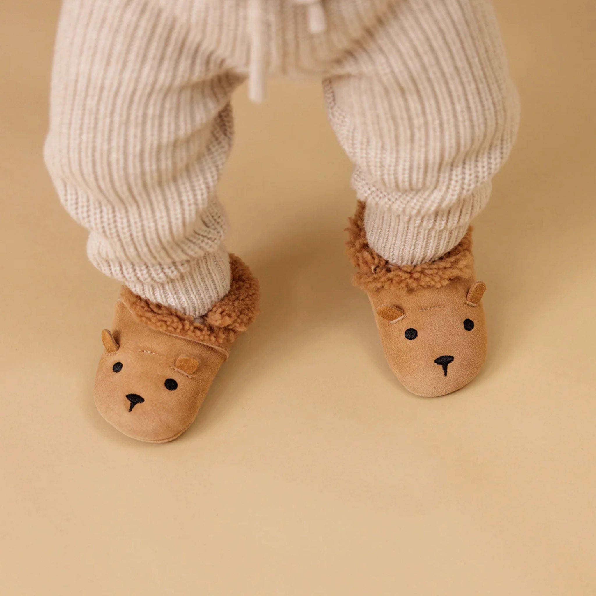 Children's bear-themed slippers worn with ribbed pants on a beige background