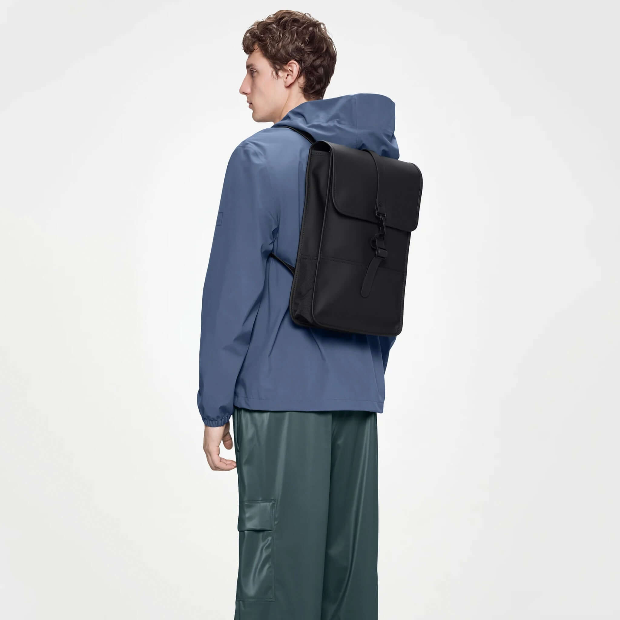 A person wearing a blue jacket and green pants carries the Rains Backpack Mini - Black.