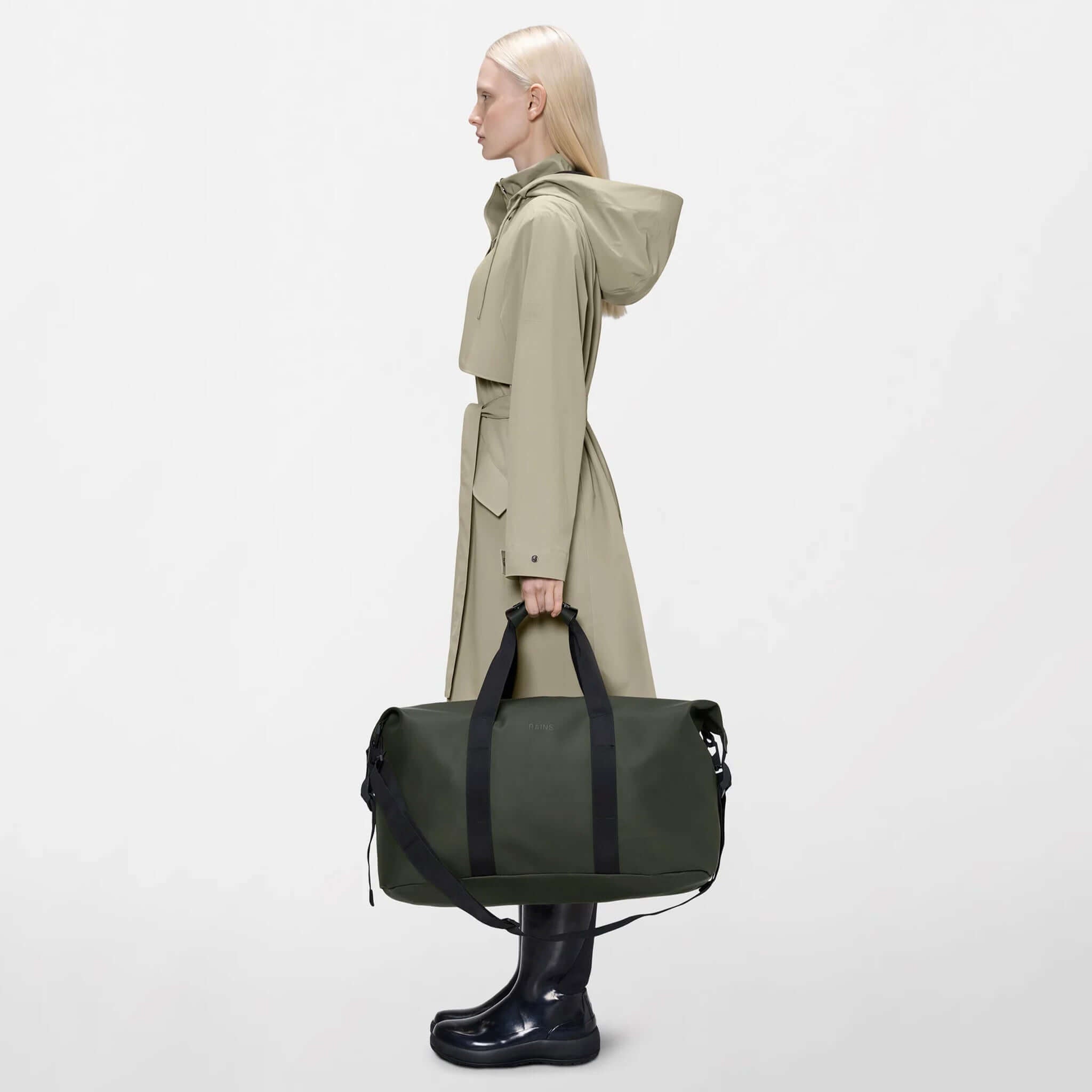 Model carrying the Rains Hilo Weekend Bag - Green while wearing a stylish raincoat.