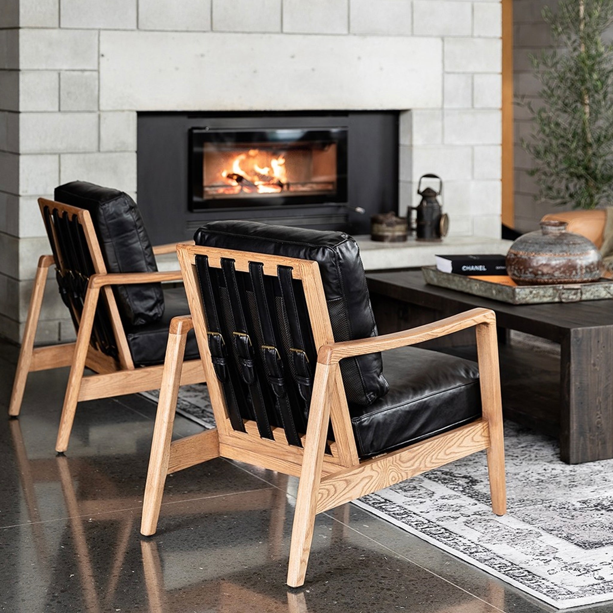 A contemporary armchair with a natural wood frame and black leather upholstery, featuring a high backrest with a brass buckle and joined canvas and leather straps.