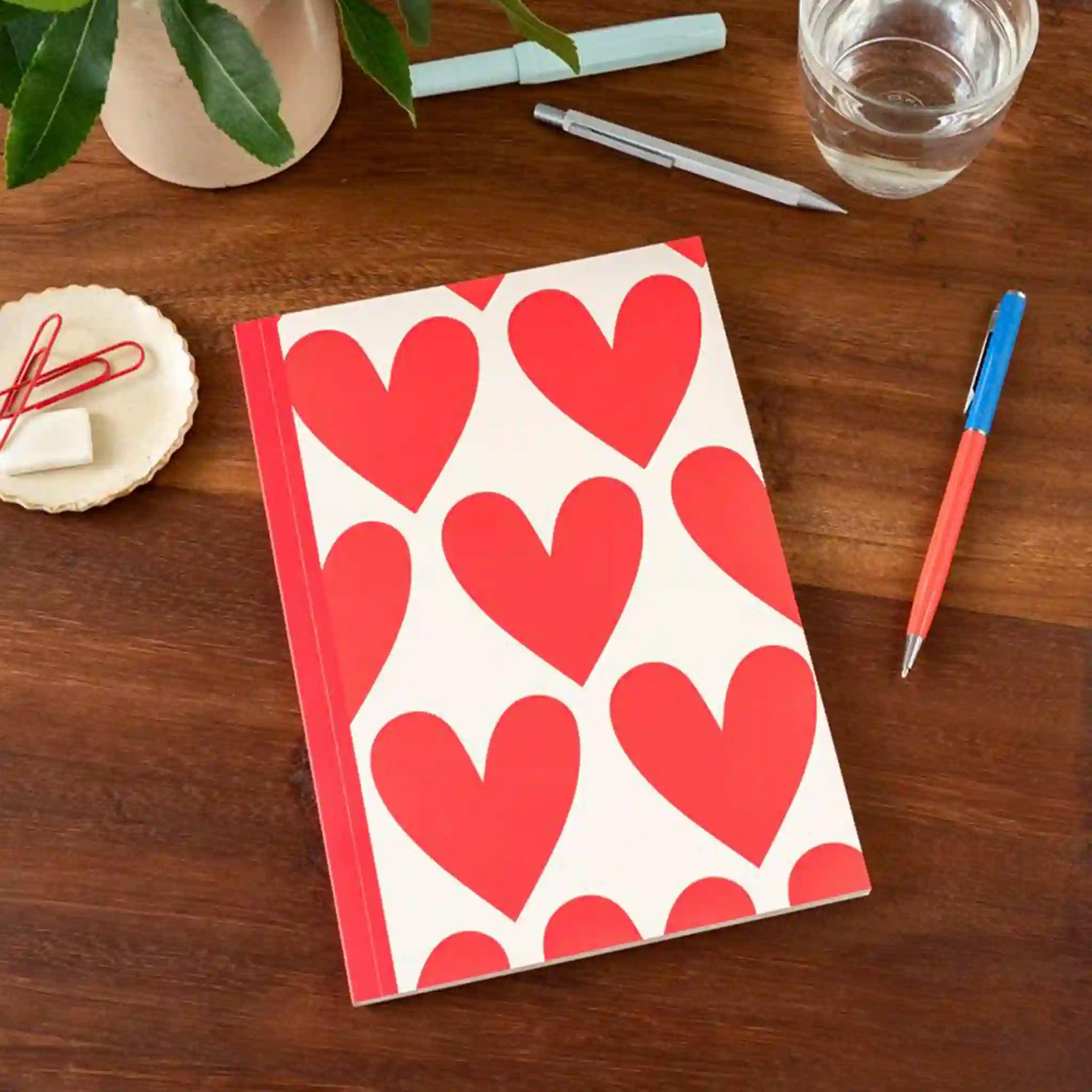 Notebook with red heart pattern on a wooden table with pens and a glass of water.