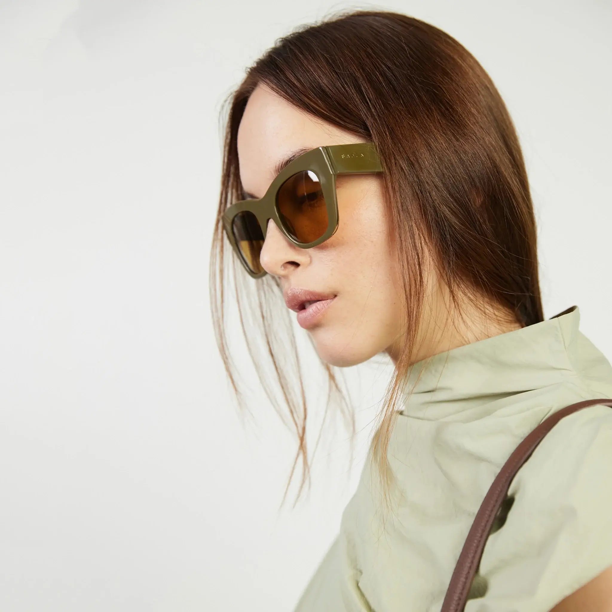 Woman wearing green sunglasses and a light-colored jacket against a plain background