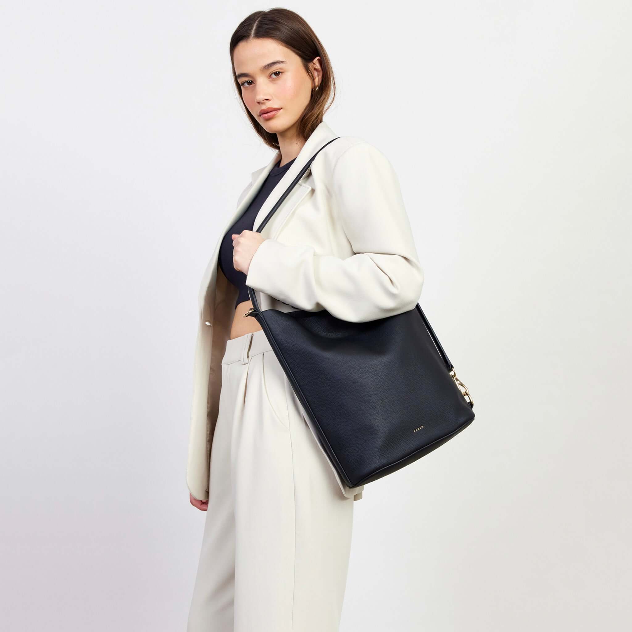 Alt Tag: Saben June Shoulder Bag - Black Luxe bucket-style tote with adjustable shoulder strap, worn crossbody on model in cream suit