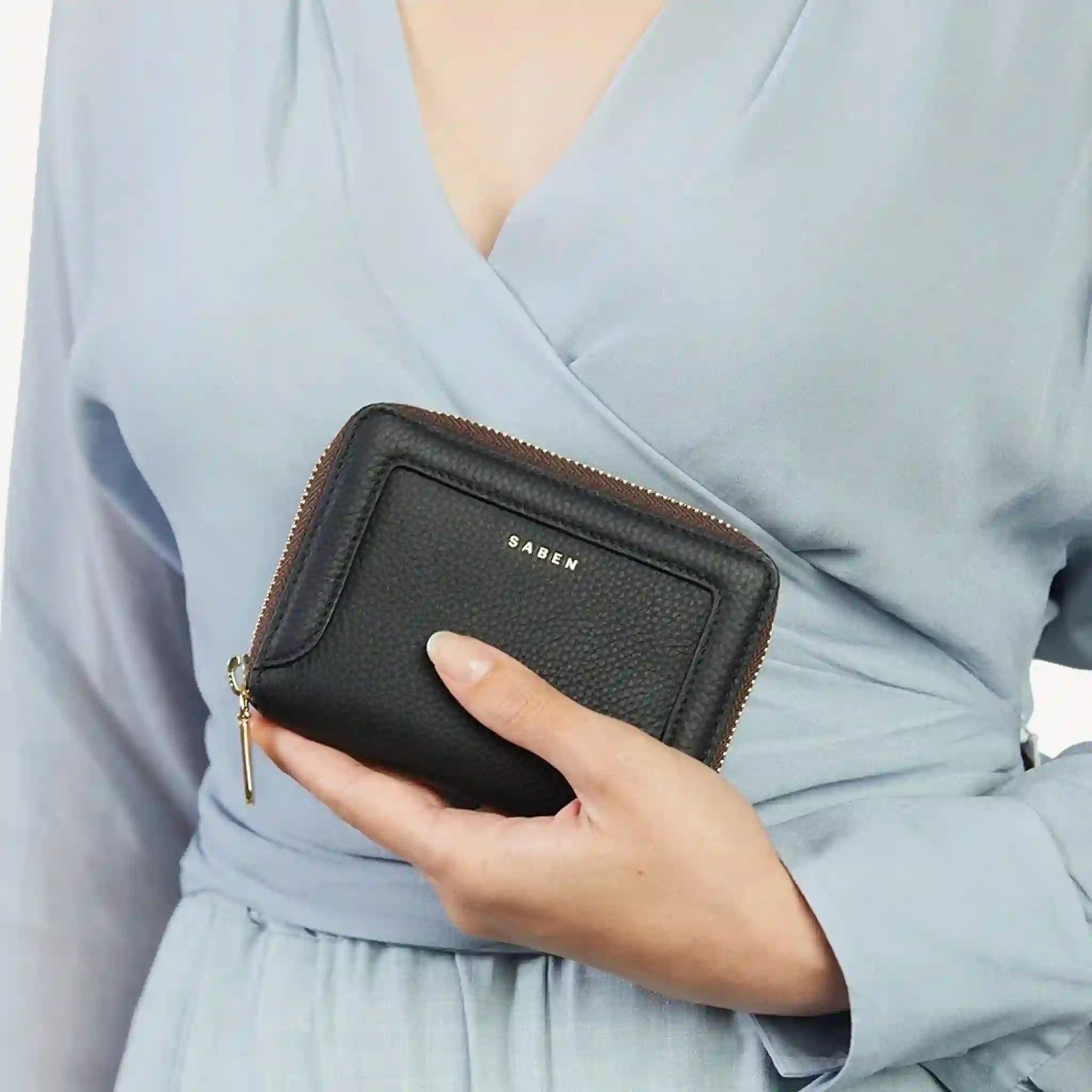 Person holding a black wallet with 'Saben' branding against a light gray background
