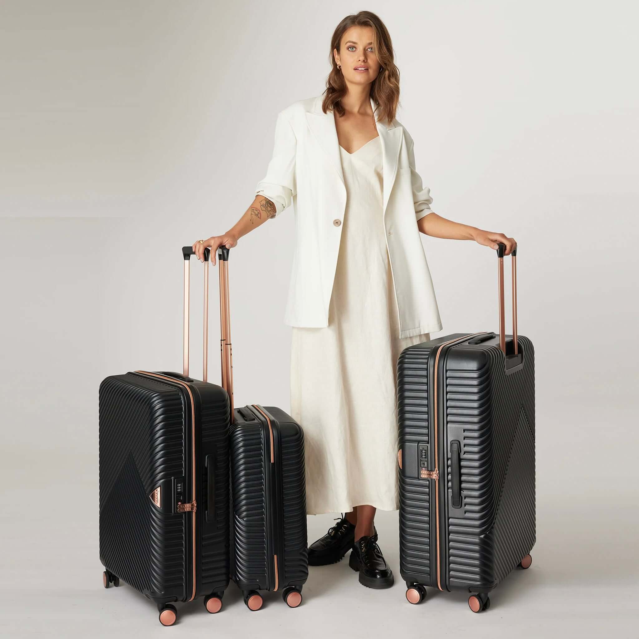 Woman with a set of three black suitcases with metallic rose gold detailing, including a cabin size, a medium size, and a large size, displayed with their handles up.
