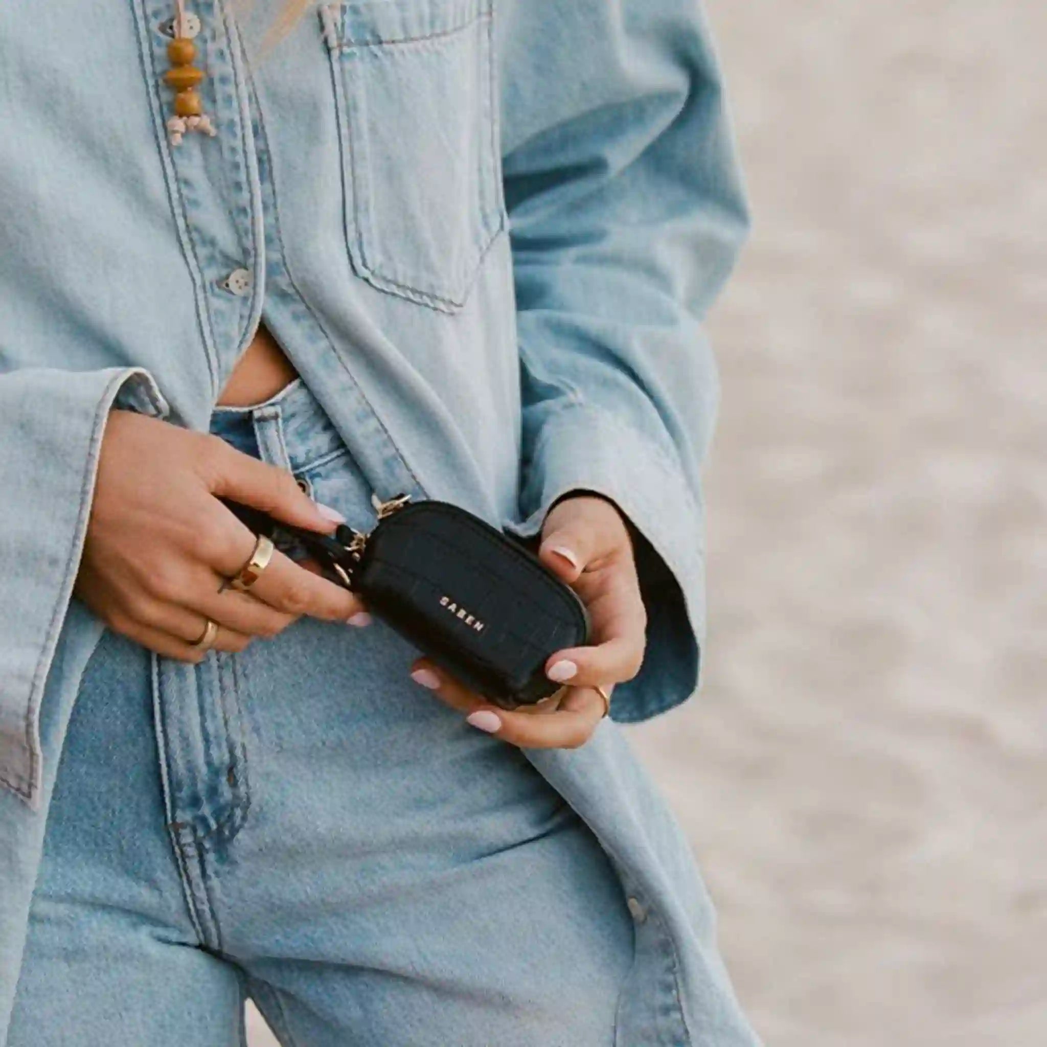 Person holding a black handbag with 'Saben' branding, wearing a light blue denim jacket and jeans.