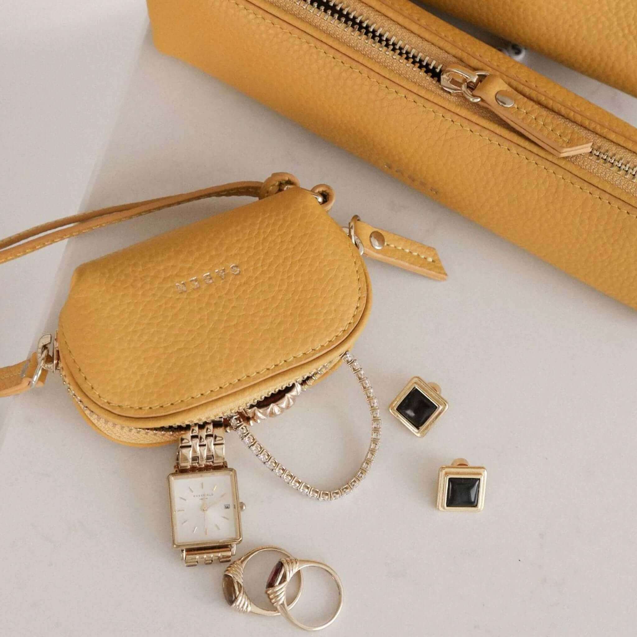 Yellow handbag with accessories including a watch, rings, and earrings on a light background