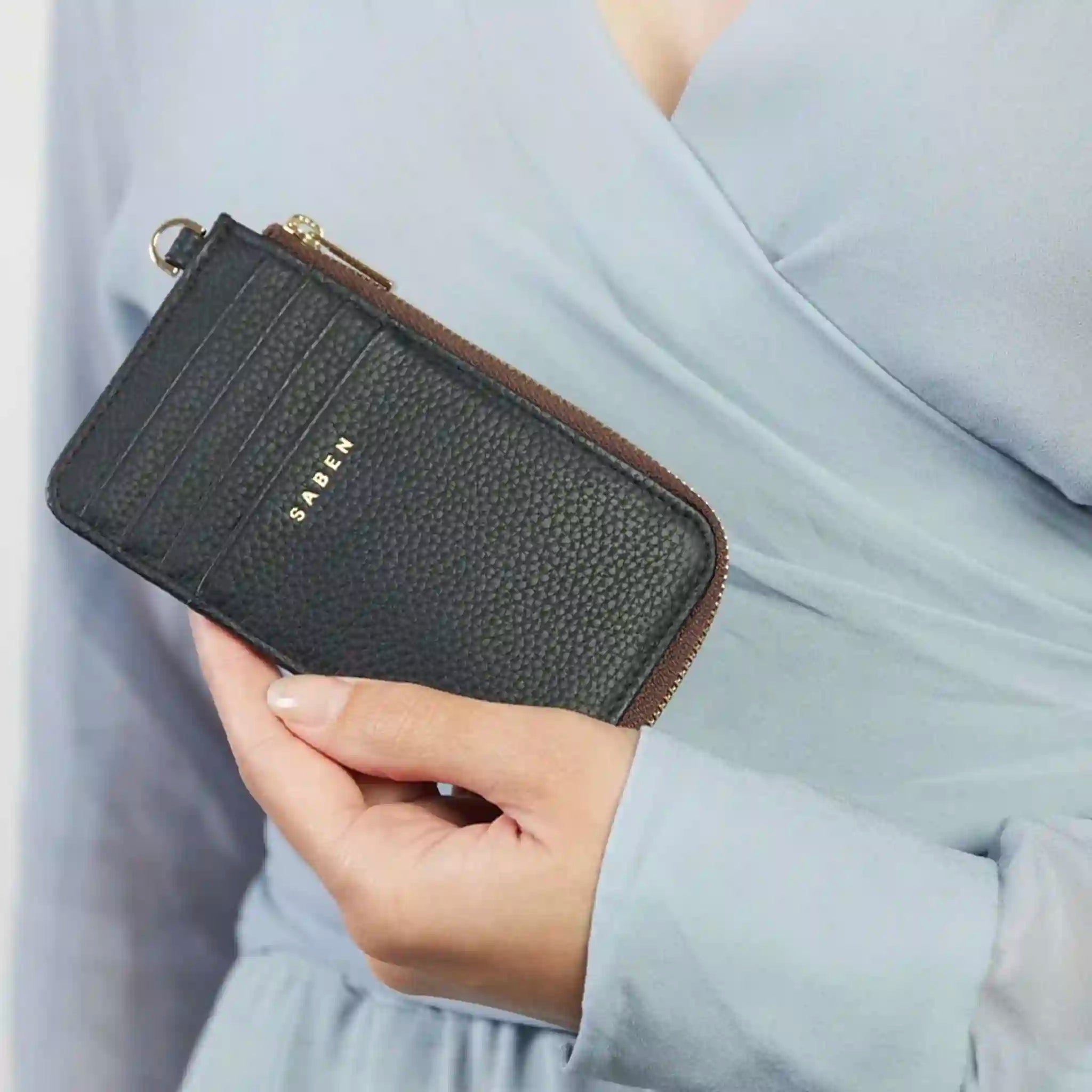 Person holding a black wallet with 'SABEN' branding against a light gray background