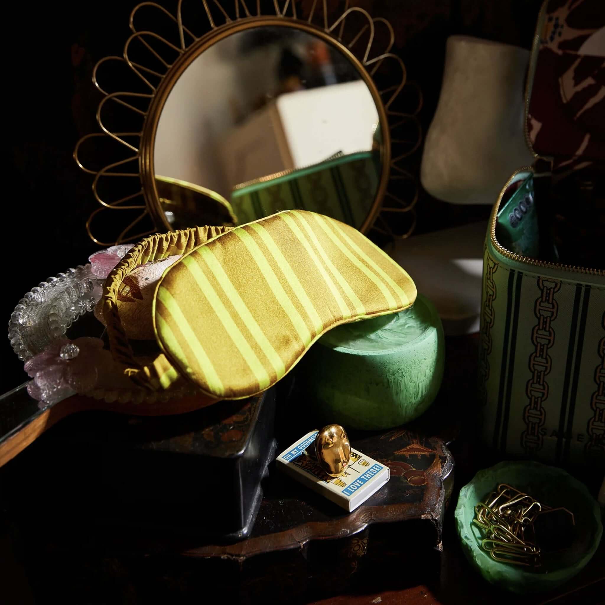 An olive green silk eye mask laid out on a surface with various other items, such as a mirror reflecting a green wall, a green candle holder, and a small container.