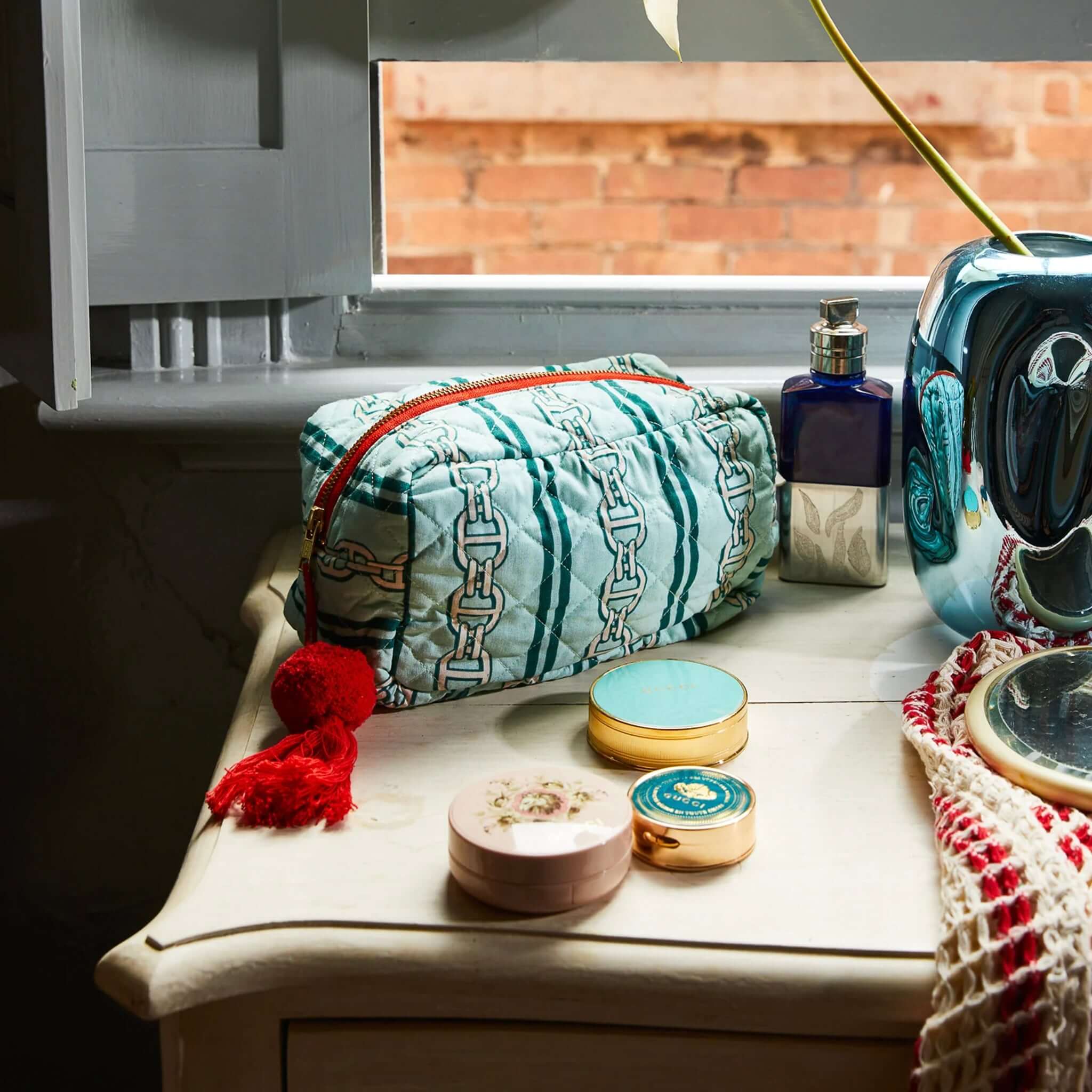 A green and multicolored striped beauty bag with a quilted texture, orange zipper, and a red tassel on a table.