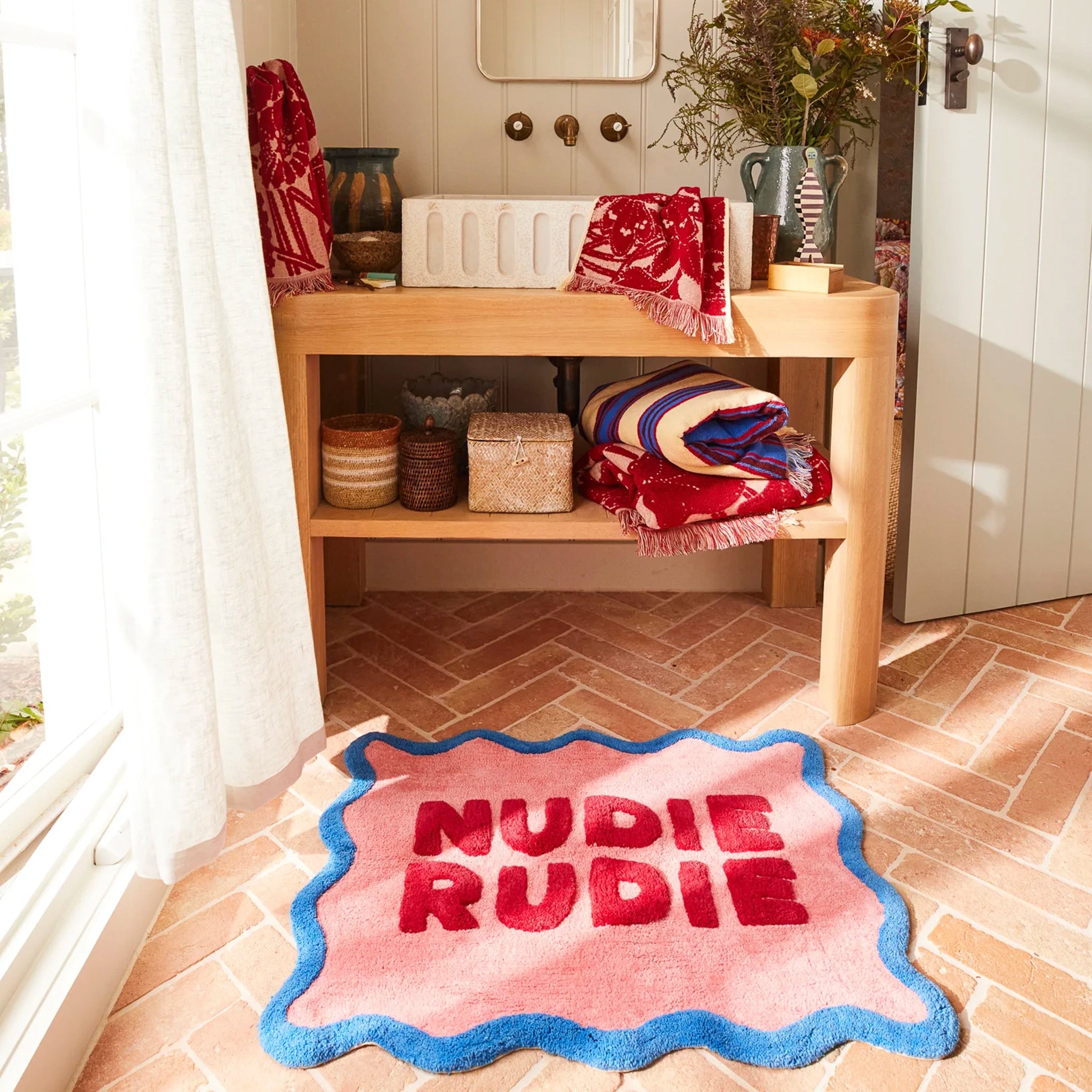 Bathroom with wooden shelf, towels, and a pink rug with 'Nudie Rudie' text.