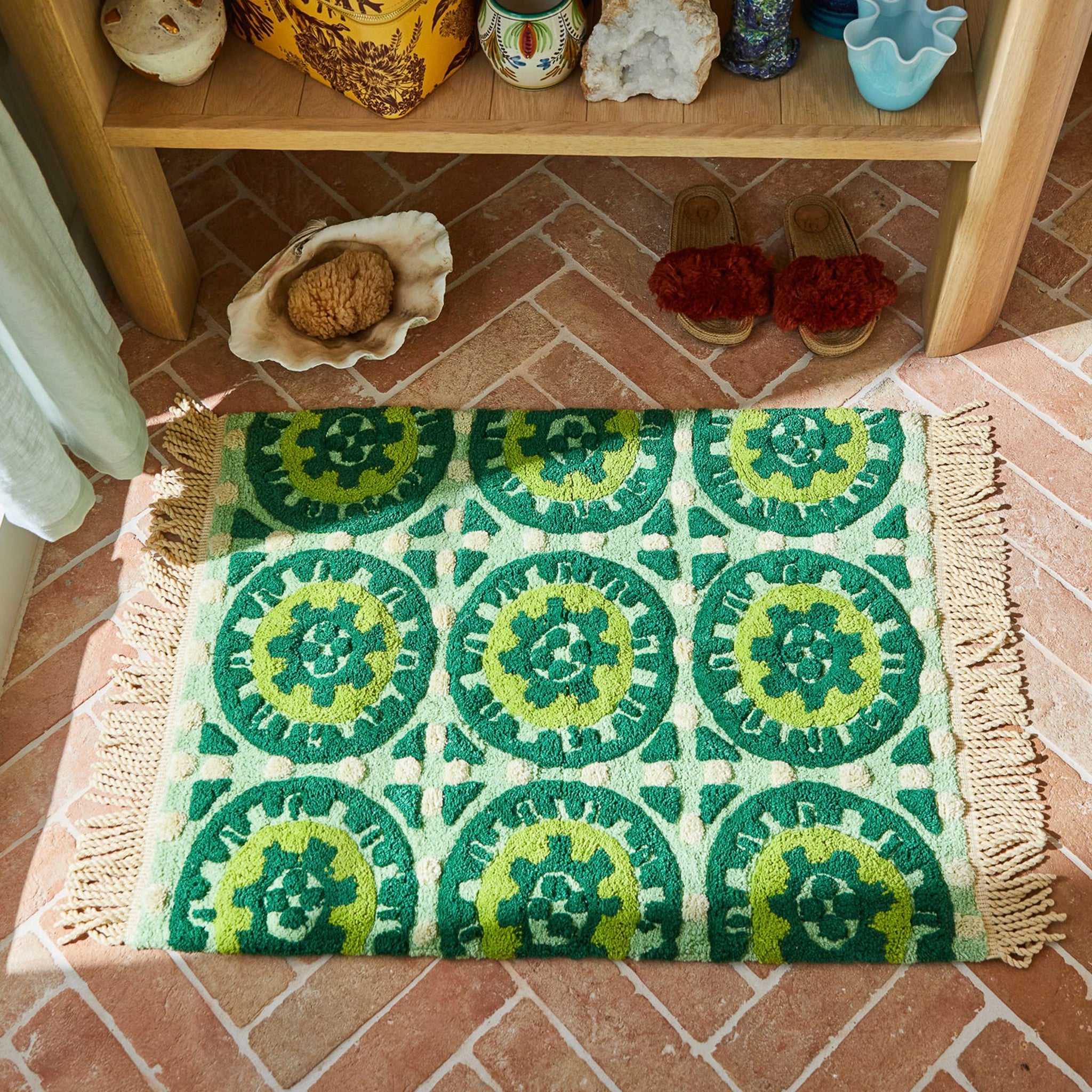 Green and yellow patterned bath mat with tassels on a brick floor, with a wooden shelf in the background.