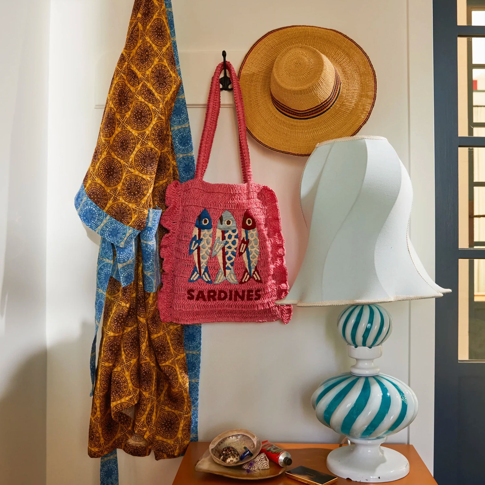 Decorative scene with a pink bag, straw hat, and scarf hanging on a hook, next to a striped lamp.