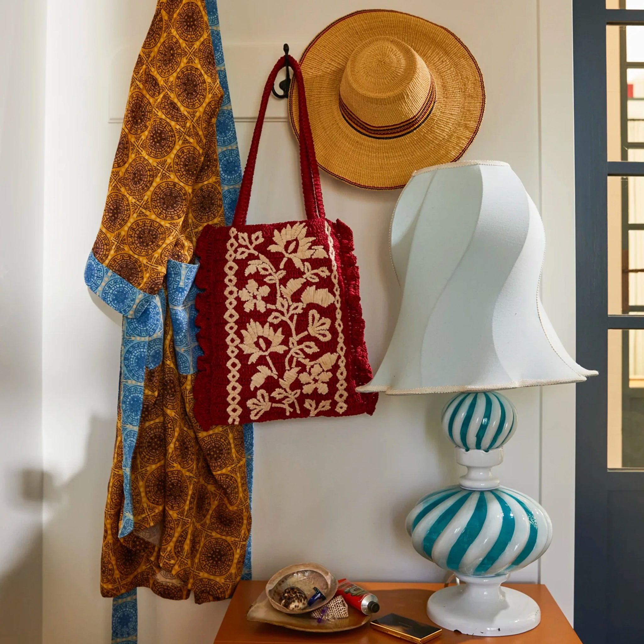 Decorative lamp with striped design on a wooden surface, next to a red bag, straw hat, and patterned towel against a white wall.