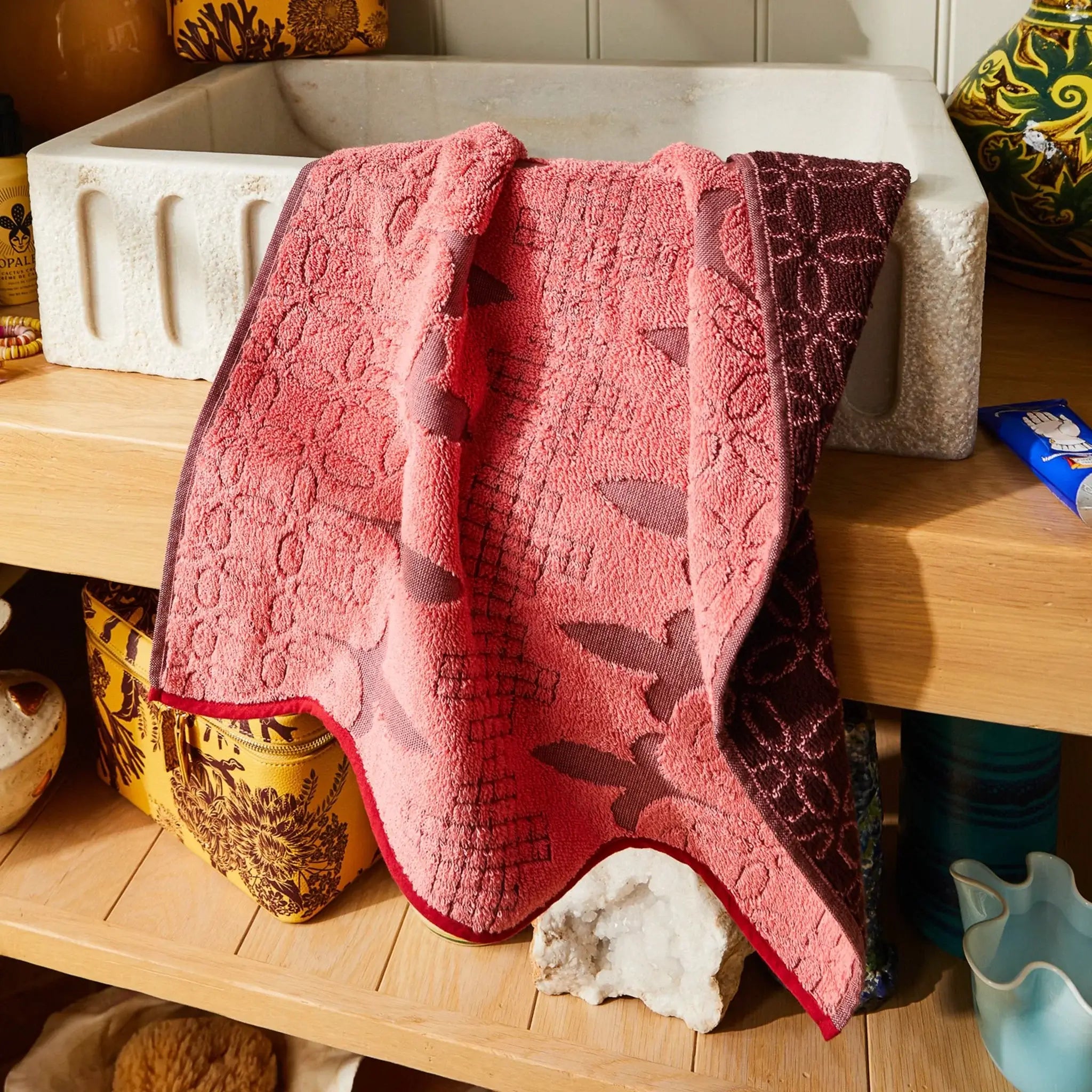 Pink textured towel draped over a wooden surface with a white container and decorative items in the background.