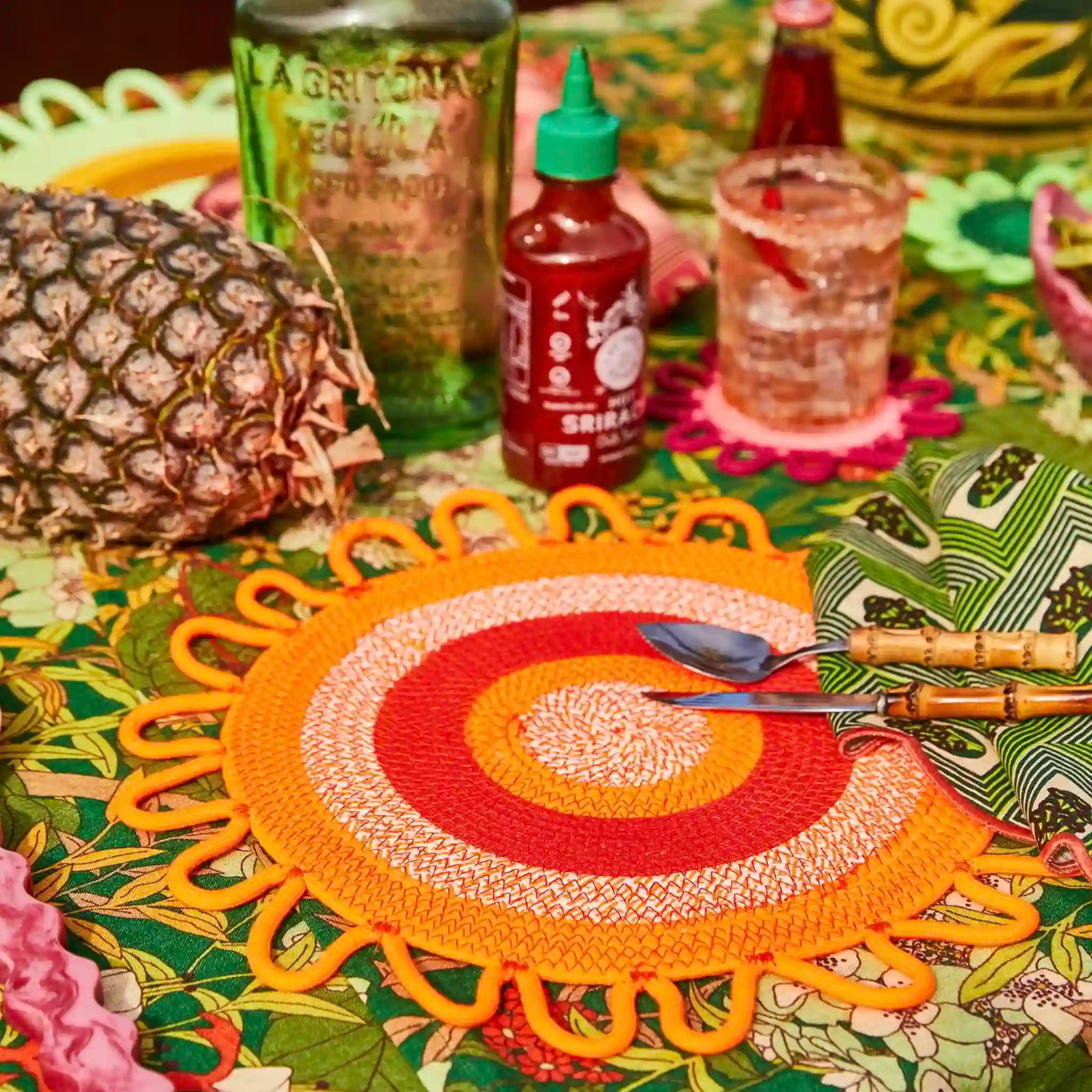 Colorful placemat on a table with a pineapple, bottle, and glass.